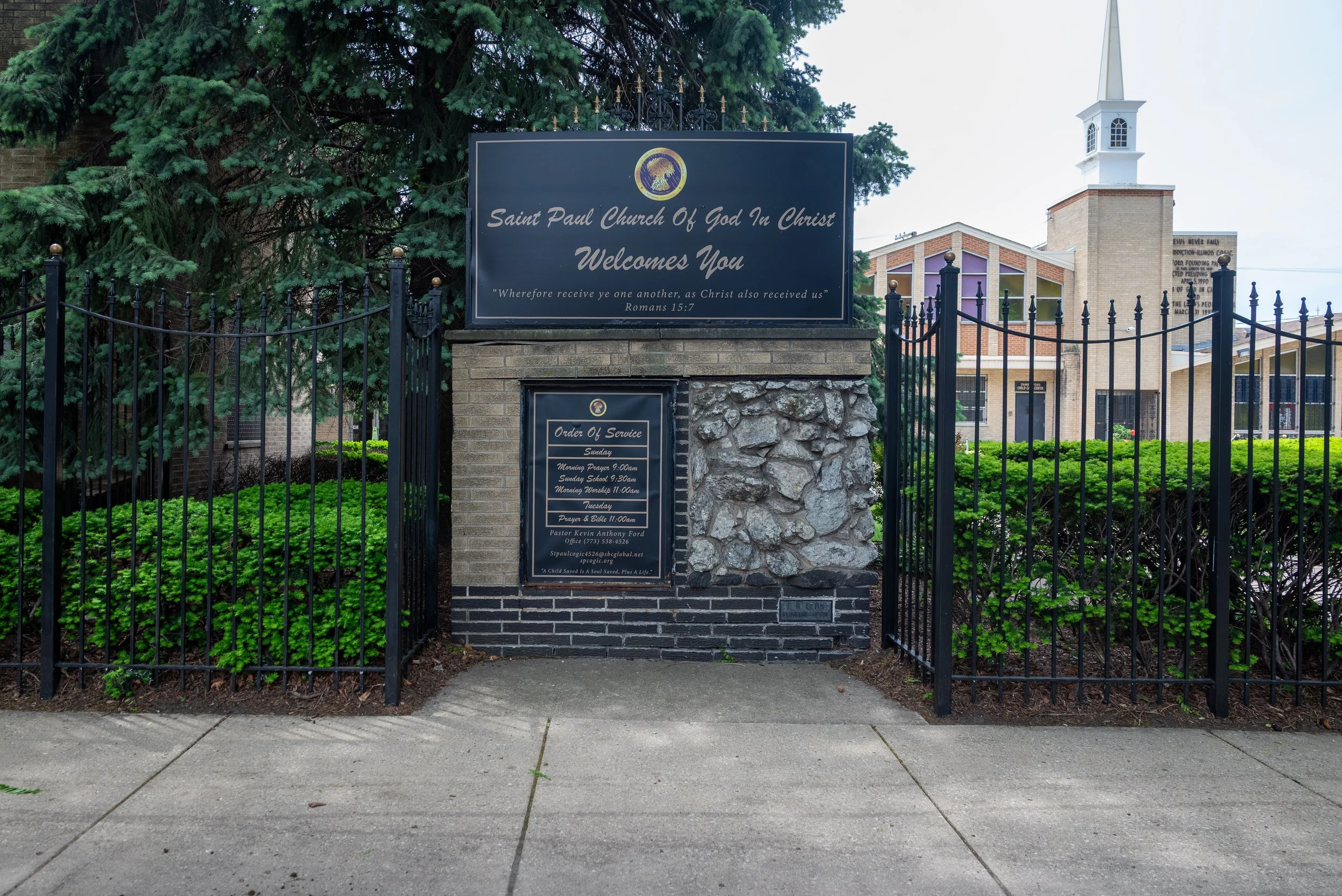 Entrance gate to Saint Paul Church of God in Christ, with a large black sign displaying the church's name, a Bible verse from Romans 15:7, and service schedule. The church building with a white steeple is visible in the background, surrounded by gree