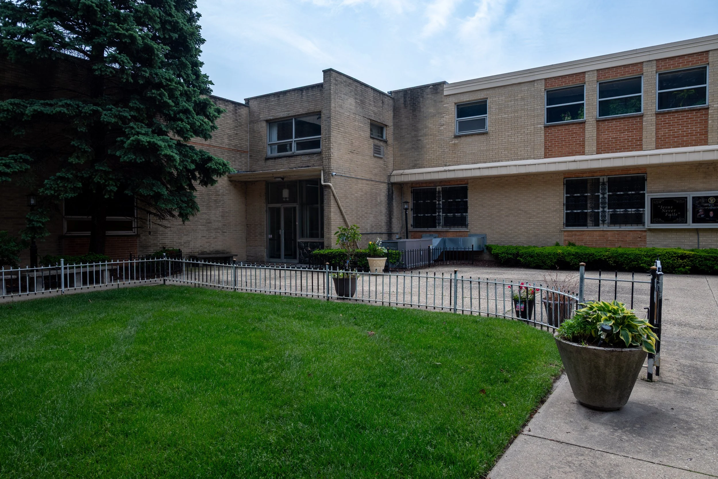 A large brick building with multiple windows and an entrance, surrounded by a well-maintained lawn, with potted plants and a small fenced area in front, and a large evergreen tree on the left side of the image.