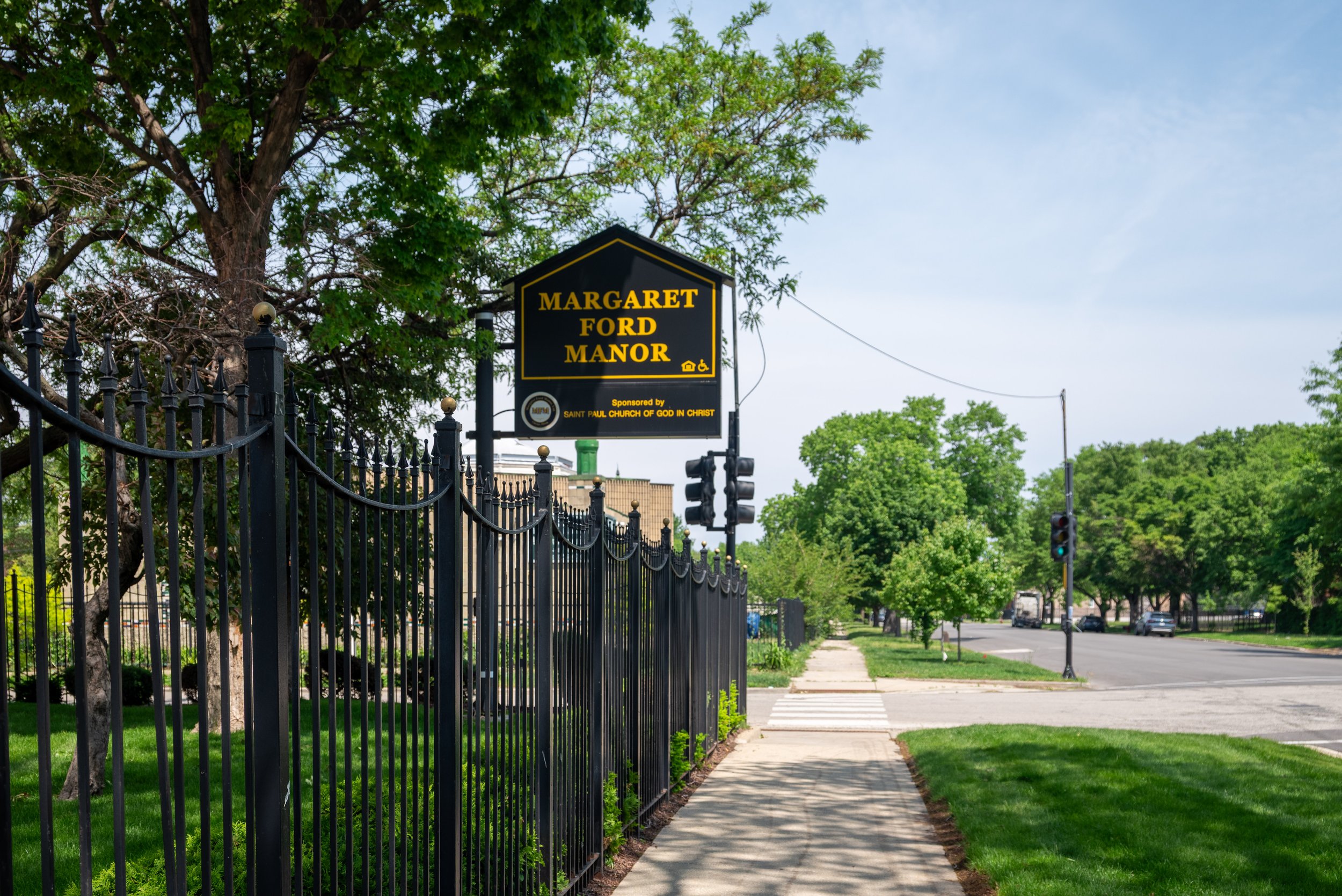 A street view with a black iron fence, a sidewalk, and green trees. There is a sign that reads 'Margaret Ford Manor' attached to the fence, sponsored by the Saint Paul Church of God in Christ.