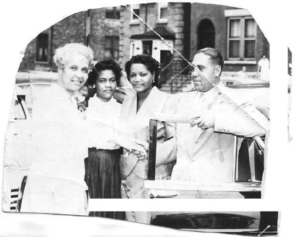 A black-and-white photo of four people standing outside a building, including two women and a man, with two young girls. They are smiling and appear to be engaged in conversation.