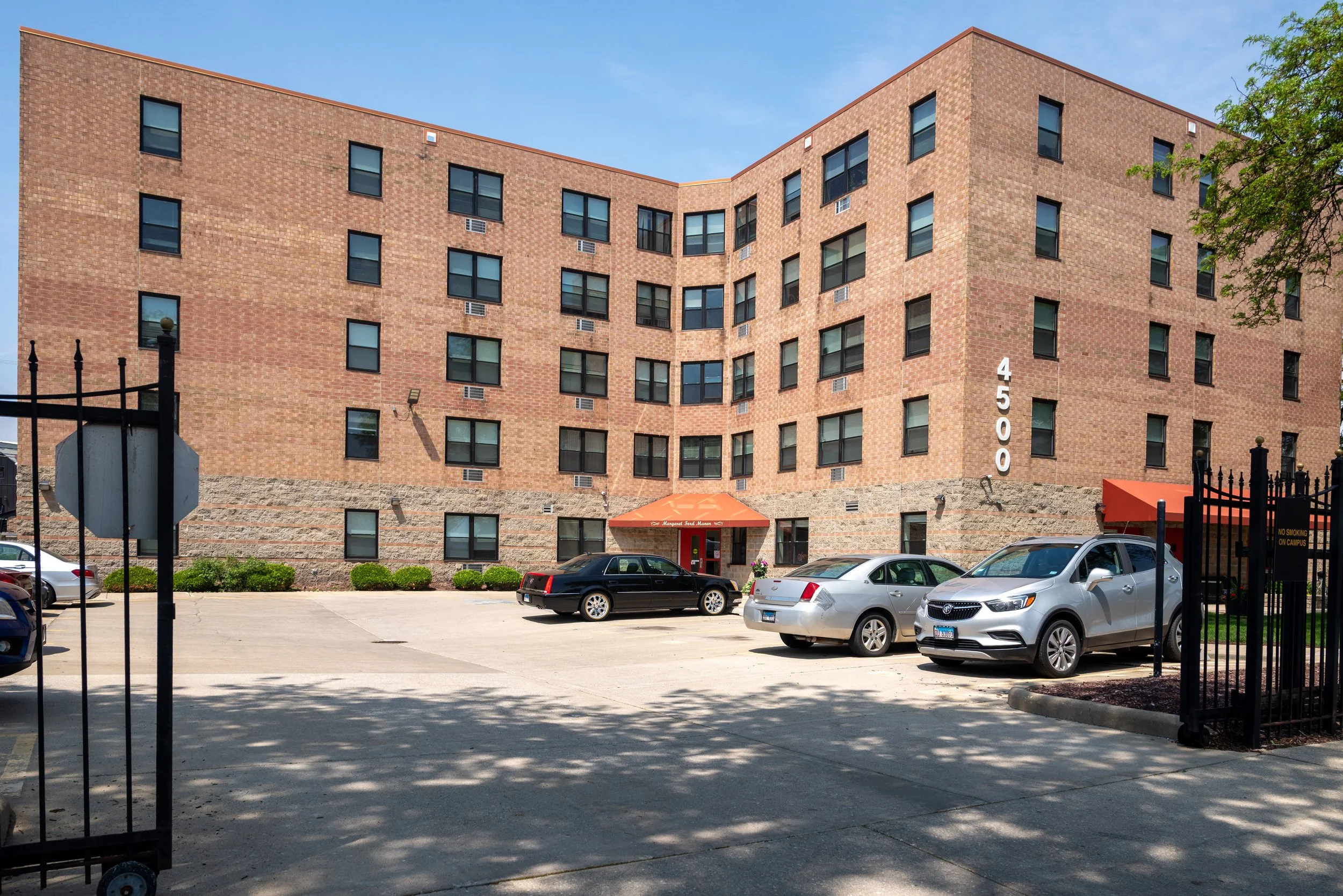 A multi-story brick building with many windows and the number 4500 displayed vertically on the right side. There is a parking lot in front with several cars parked, and trees and a fence on the sides. The sky is clear and blue.