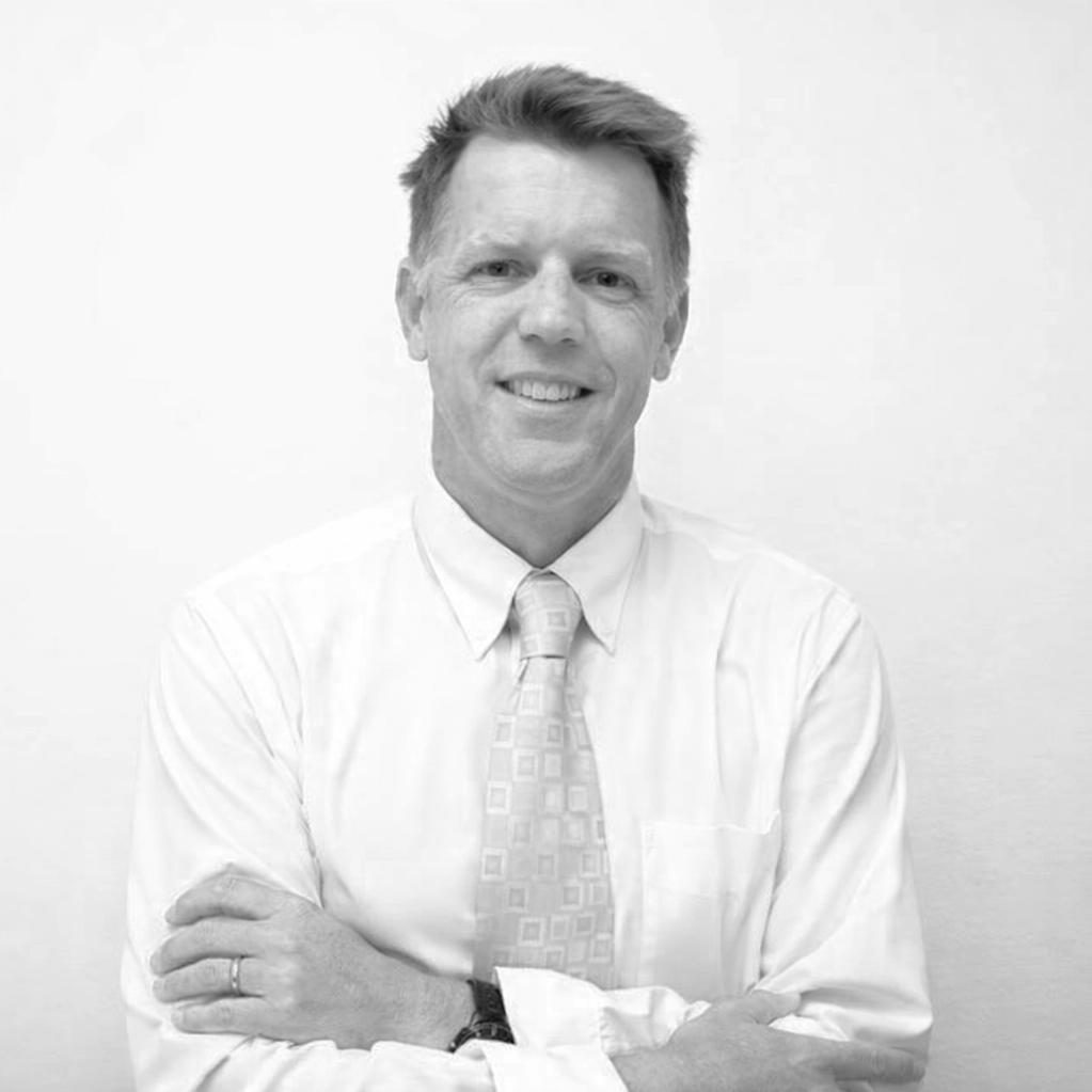 Black and white portrait of a smiling man in a long-sleeved dress shirt and tie, crossing his arms, against a plain background.