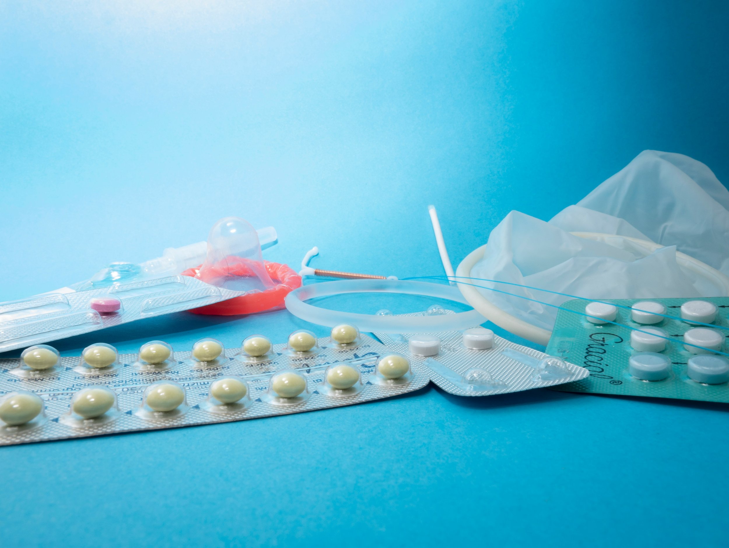 Assortment of contraceptive methods on blue background including birth control pills in blister packs, copper IUD, vaginal ring, female condom, and oral contraceptive tablet