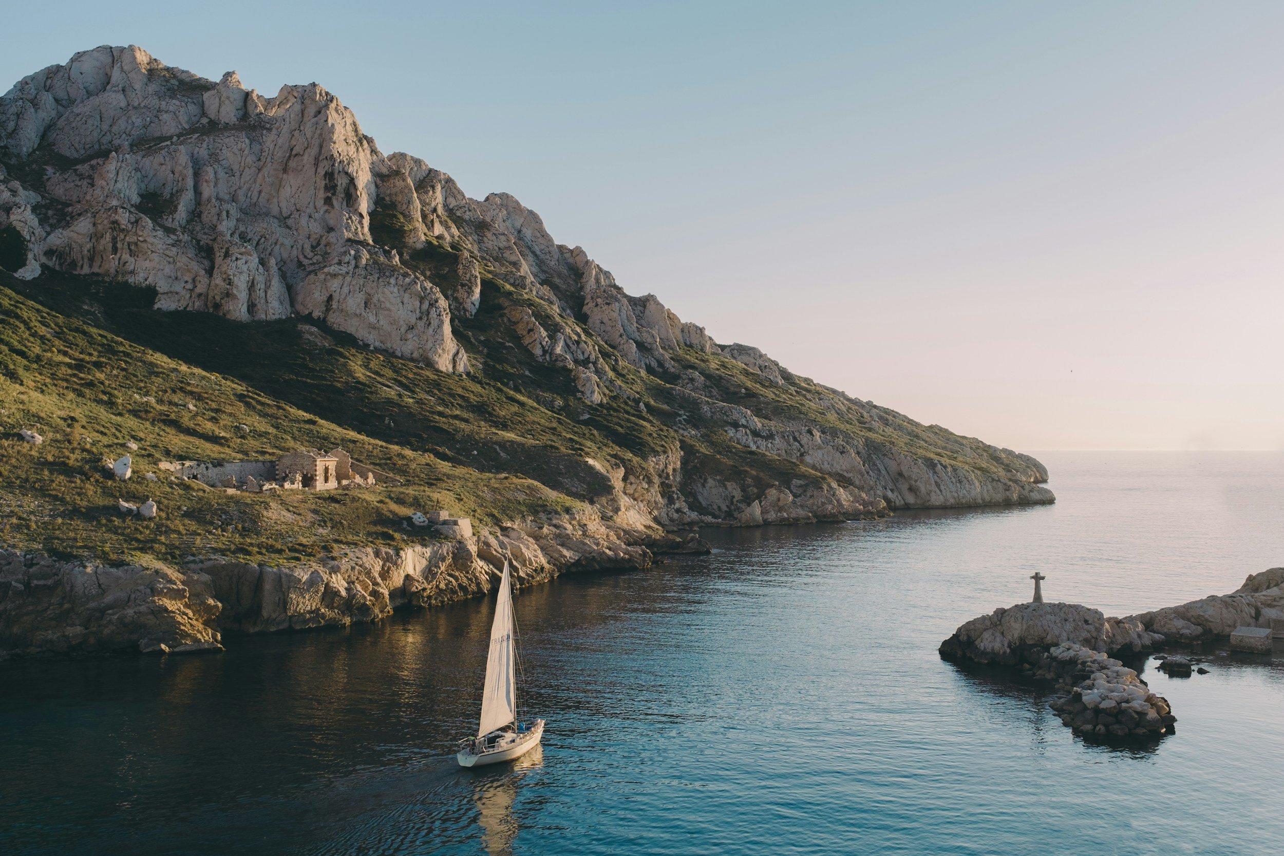Sailboat on calm water near rocky coastline with green hillside, cliffs, and a small building, under a clear sky.