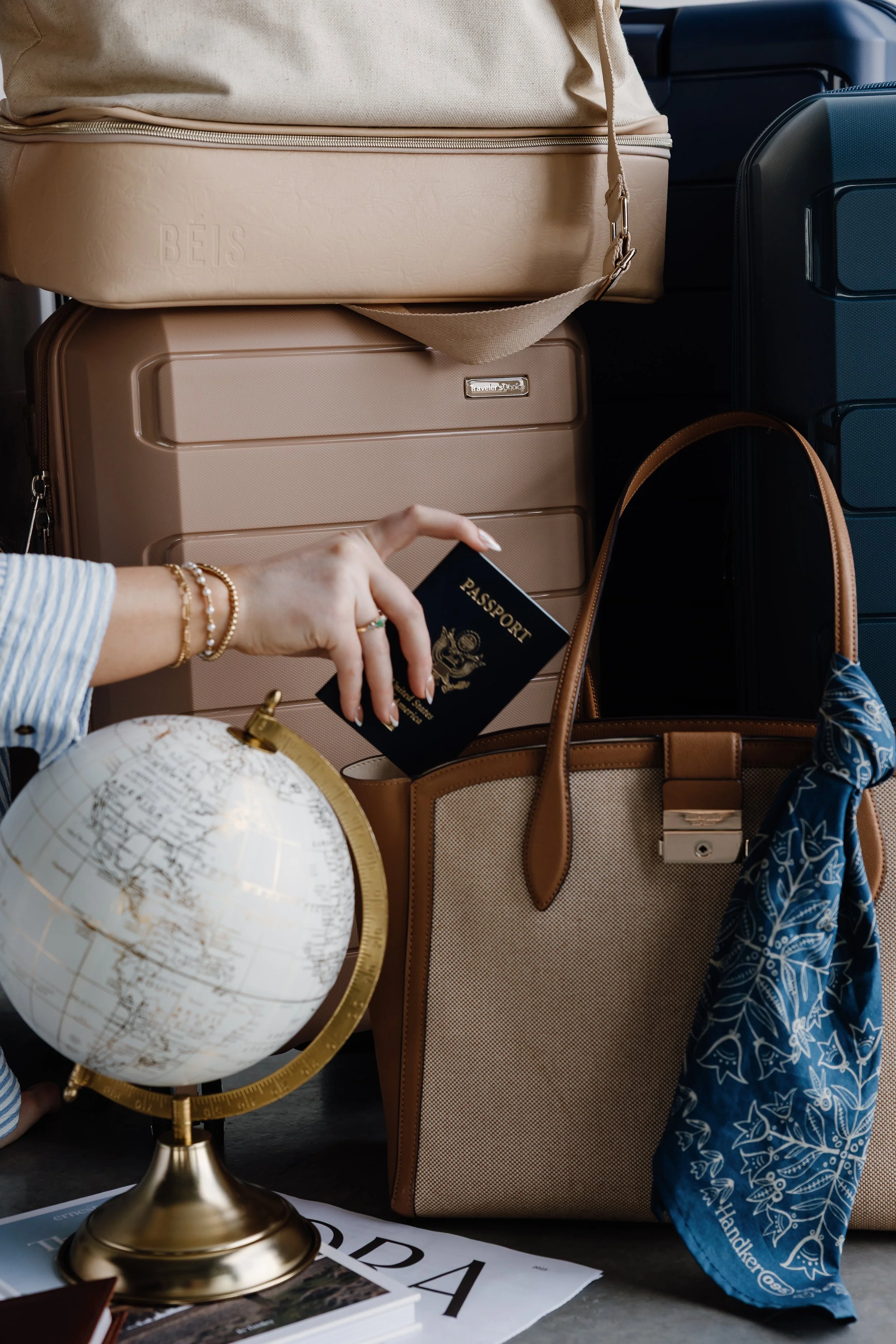 Travel essentials including a beige suitcase, a tan handbag, a person's hand holding a passport, a globe, and magazines or brochures on a surface. There are also various luggage pieces in the background.