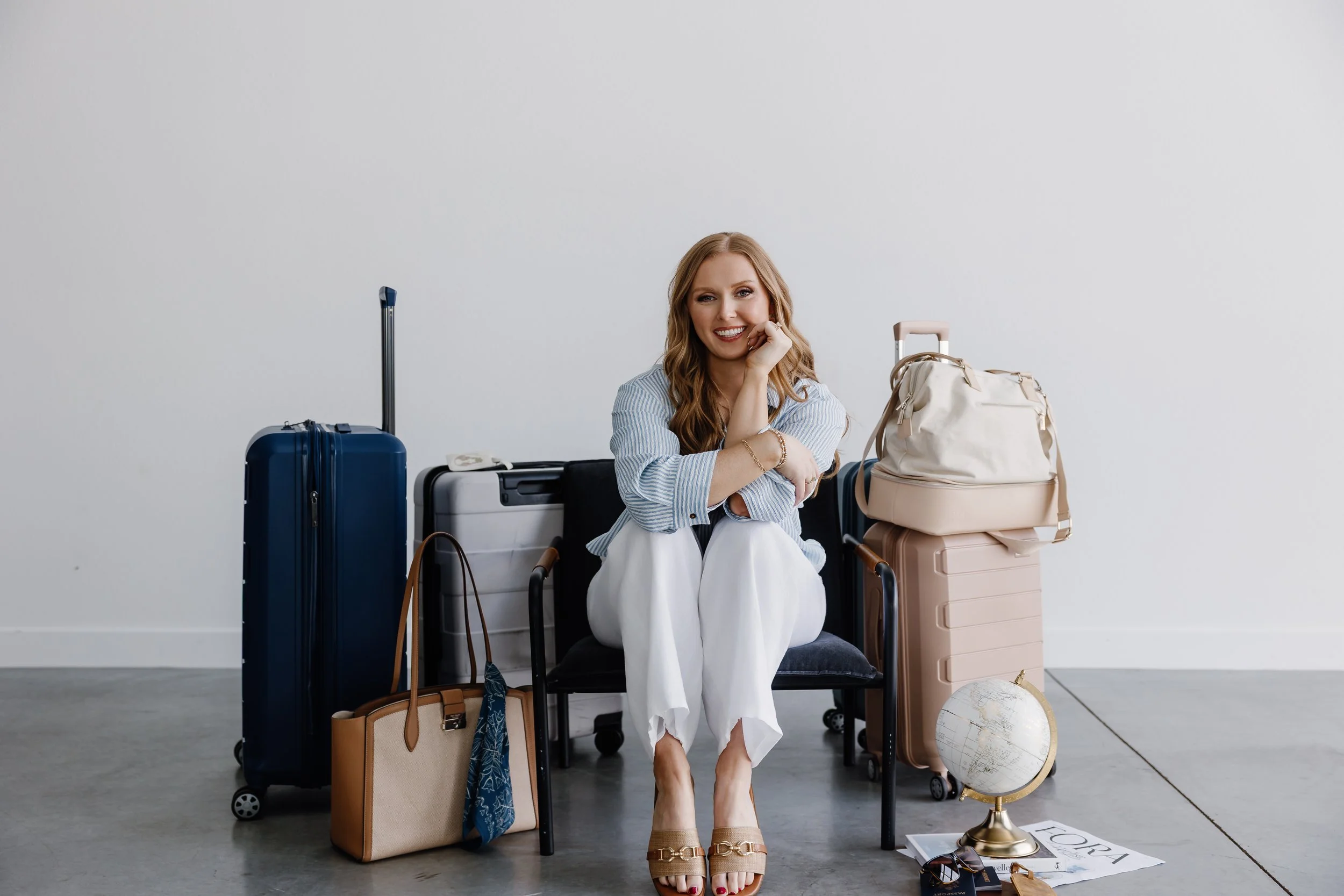 A woman with long, wavy blonde hair sitting among several suitcases and travel items in a minimalist room with a white wall. She is smiling, resting her chin on her left hand, wearing a striped blouse and white pants, with accessories and sandals. Items include a globe, a beige handbag, a world map, sunglasses, and magazines.
