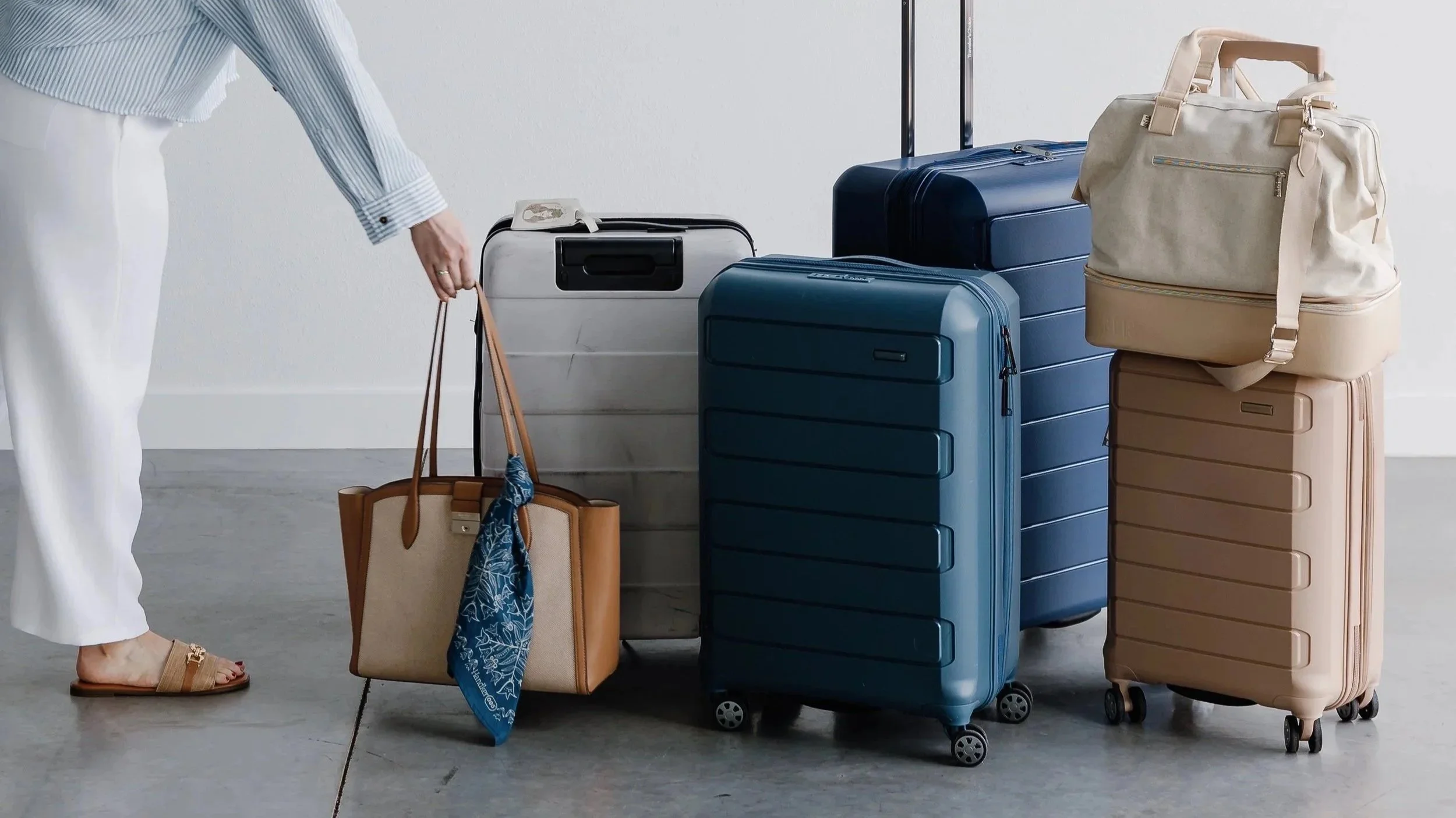 Person in white pants and striped shirt standing next to a variety of suitcases and a tote bag on a light-colored floor.
