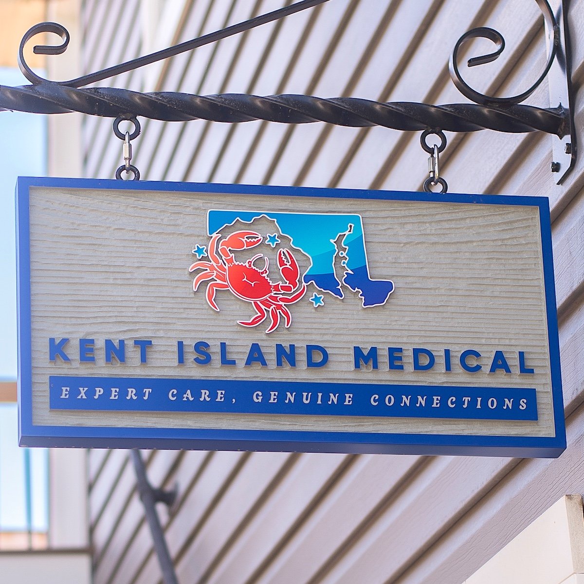 A sign hanging in front of Kent Island Medical with their name, logo, and slogan of "Expert Care, Genuine Connections"