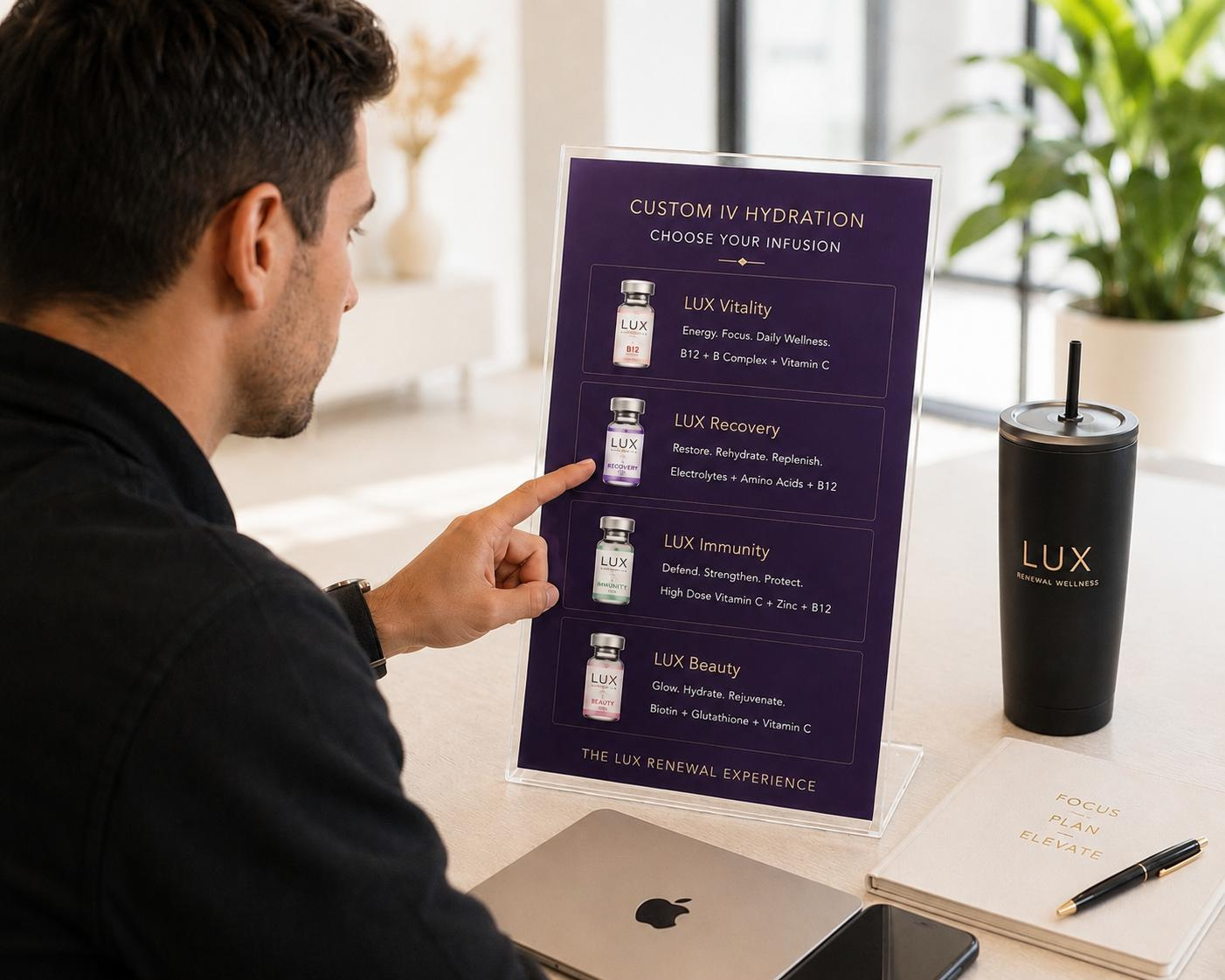 A man looking at a purple display board promoting different IV hydration infusions from LUX. The board showcases options like LUX Vitality, Recovery, Immunity, and Beauty, along with their benefits and ingredients. The desk contains a black LUX tumbler with a straw, a silver Apple MacBook, a black smartphone, and a notebook with a pen.