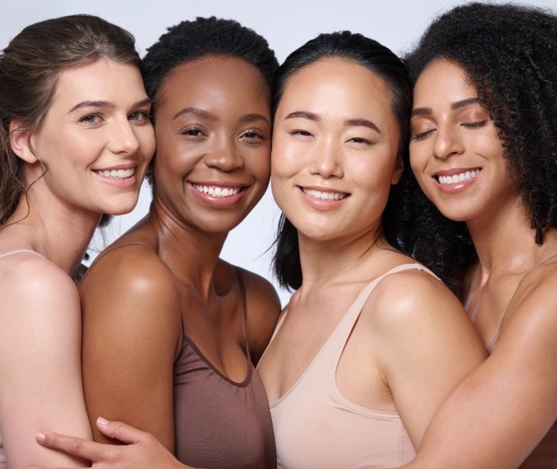 Four women standing close together, smiling, with arms around each other, against a plain, light-colored background.
