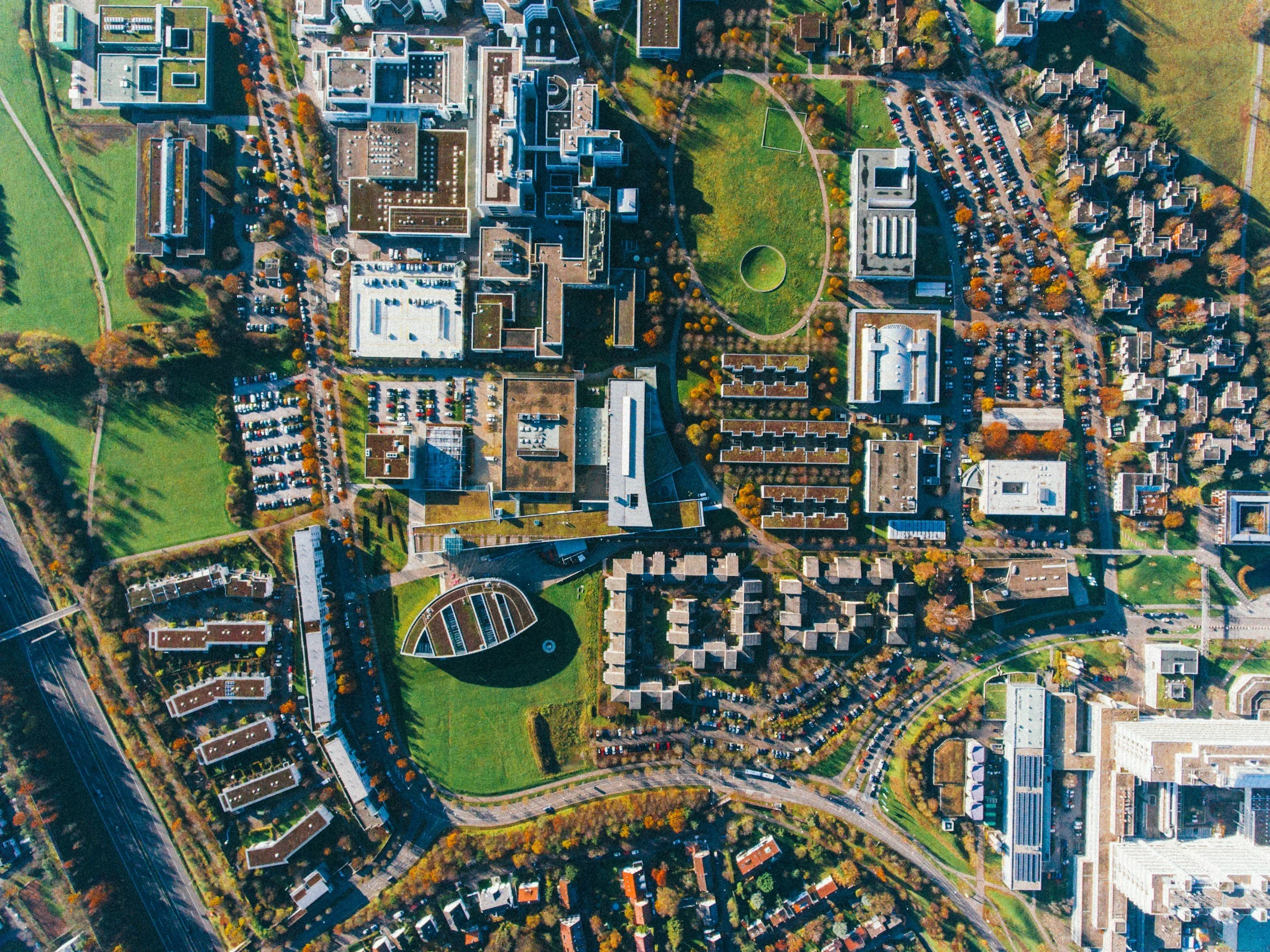 An aerial view of a cityscape with numerous buildings, parking lots, green parks, and roads in vibrant fall colors, with visible rooftops and surrounding infrastructure.