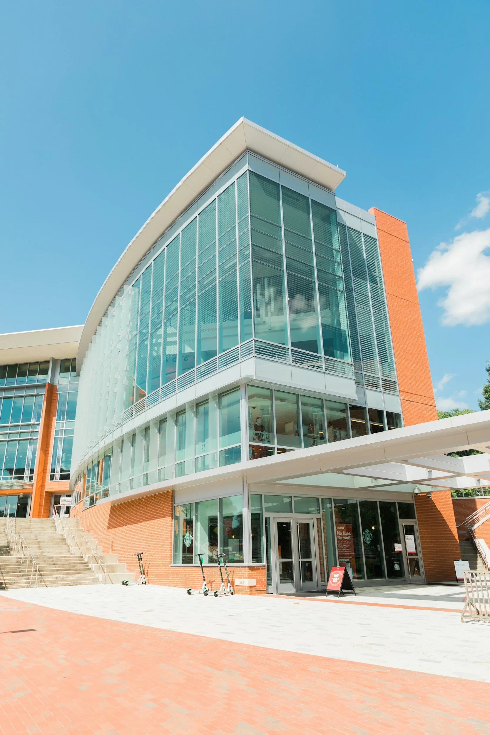 Modern building with glass windows and brick exterior, steps leading up to entrance, scooters parked outside, and a sign near the door under a clear blue sky.