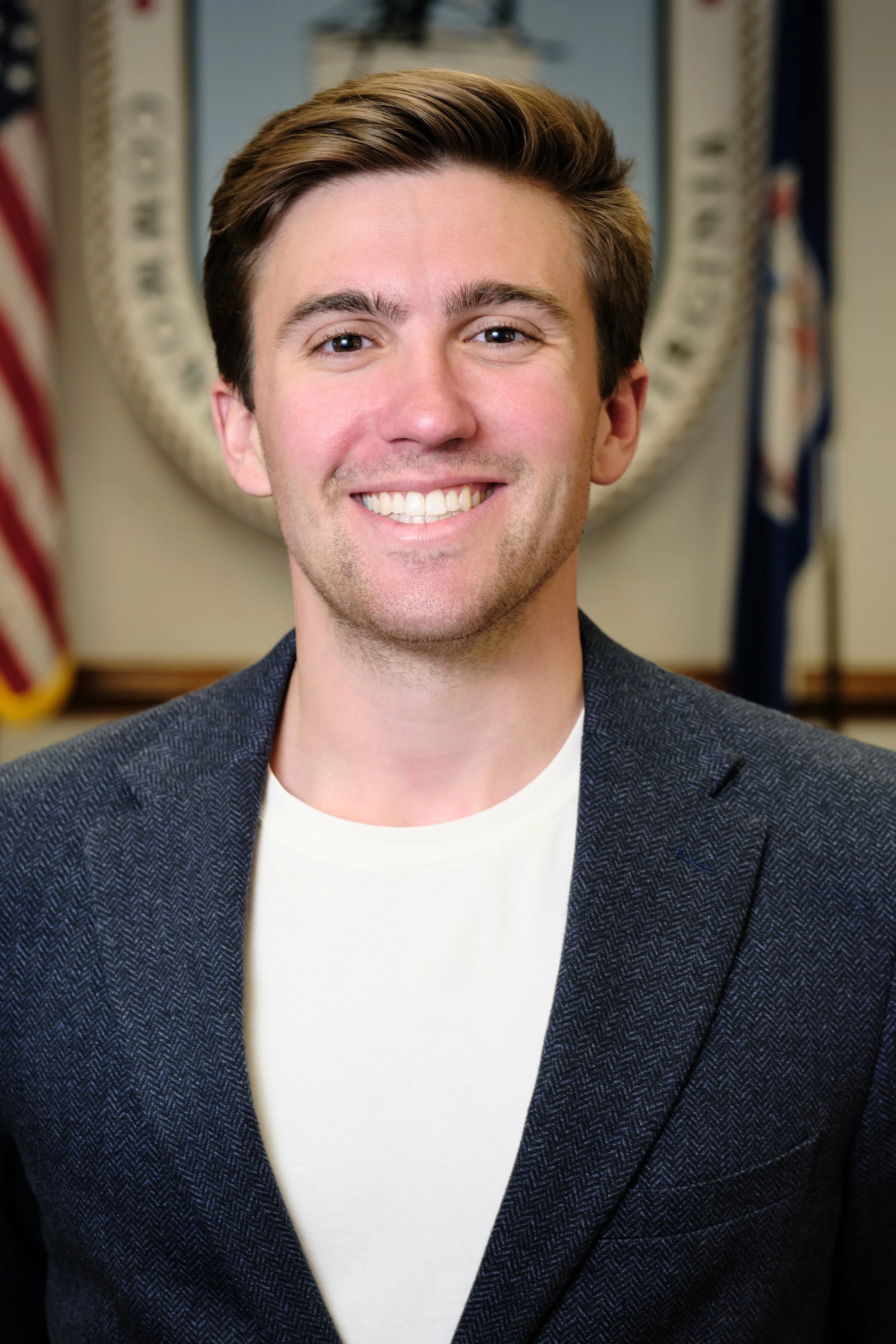 A young man with brown hair smiling, wearing a white shirt and a dark blazer, with an American flag and a government emblem in the background.