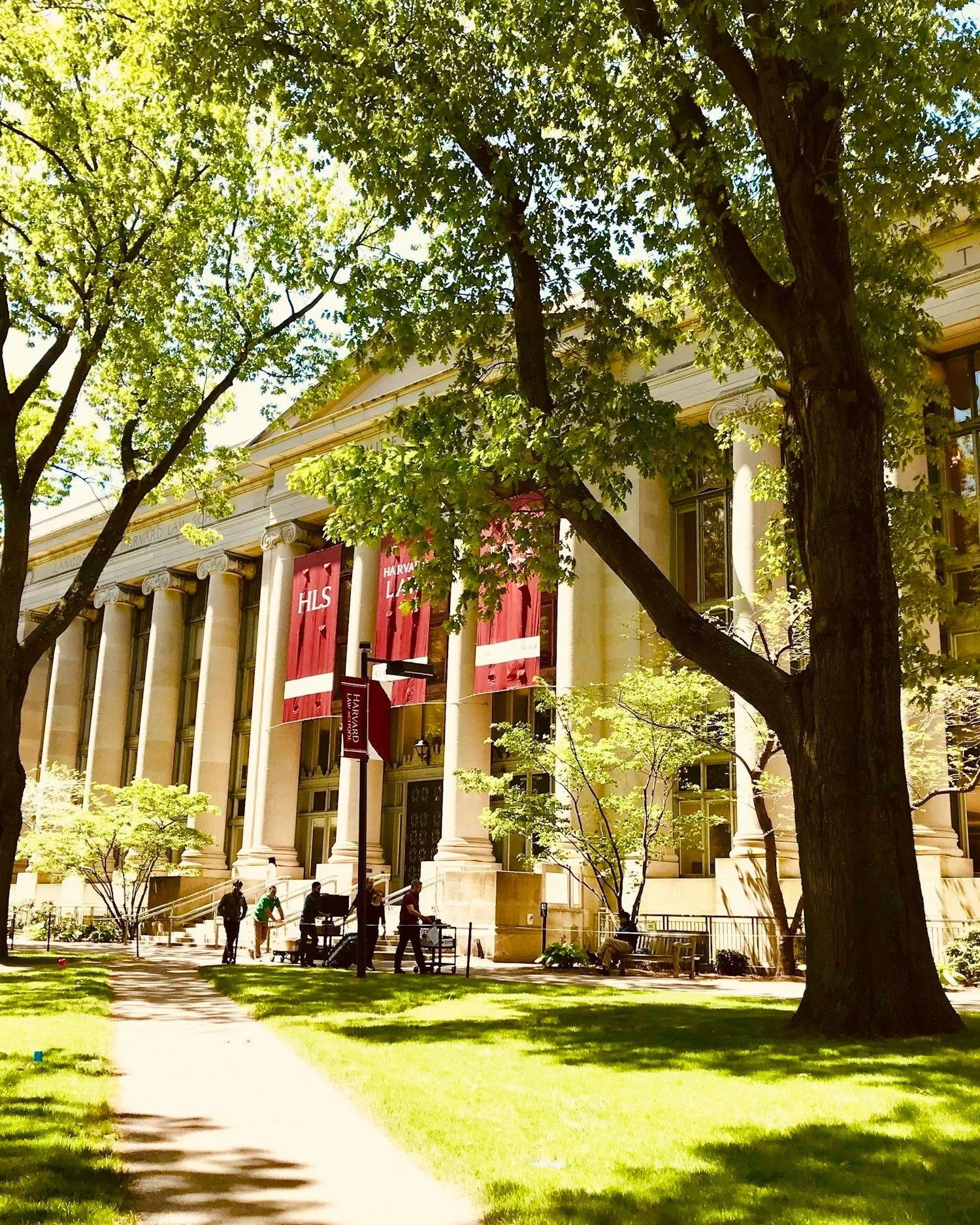 The Harvard Law School building seen through trees with green leaves, banners hanging on the columns, and people walking on the sidewalk in front.