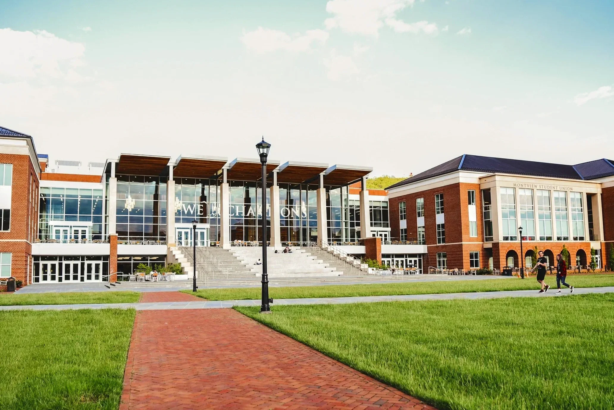 The Montview Student Union at the University of Colorado Colorado Springs, with a brick pathway leading to stairs and a glass-fronted building, green grass, street lamps, and a blue sky with clouds.