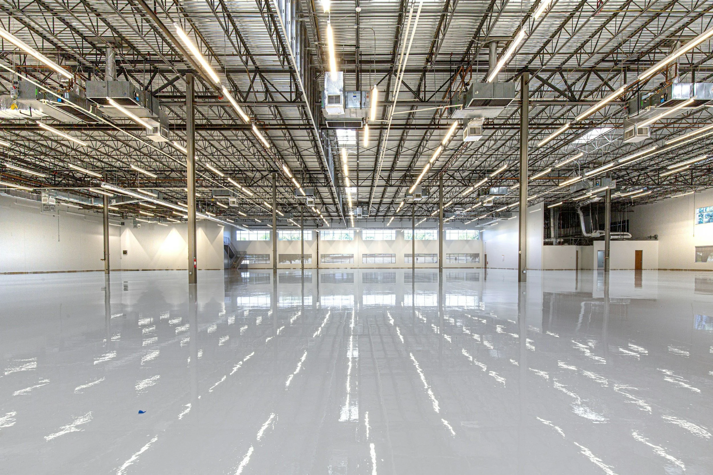 Empty industrial warehouse with high ceiling, exposed metal beams, bright lighting, and large windows, with a polished concrete floor reflecting the lights.