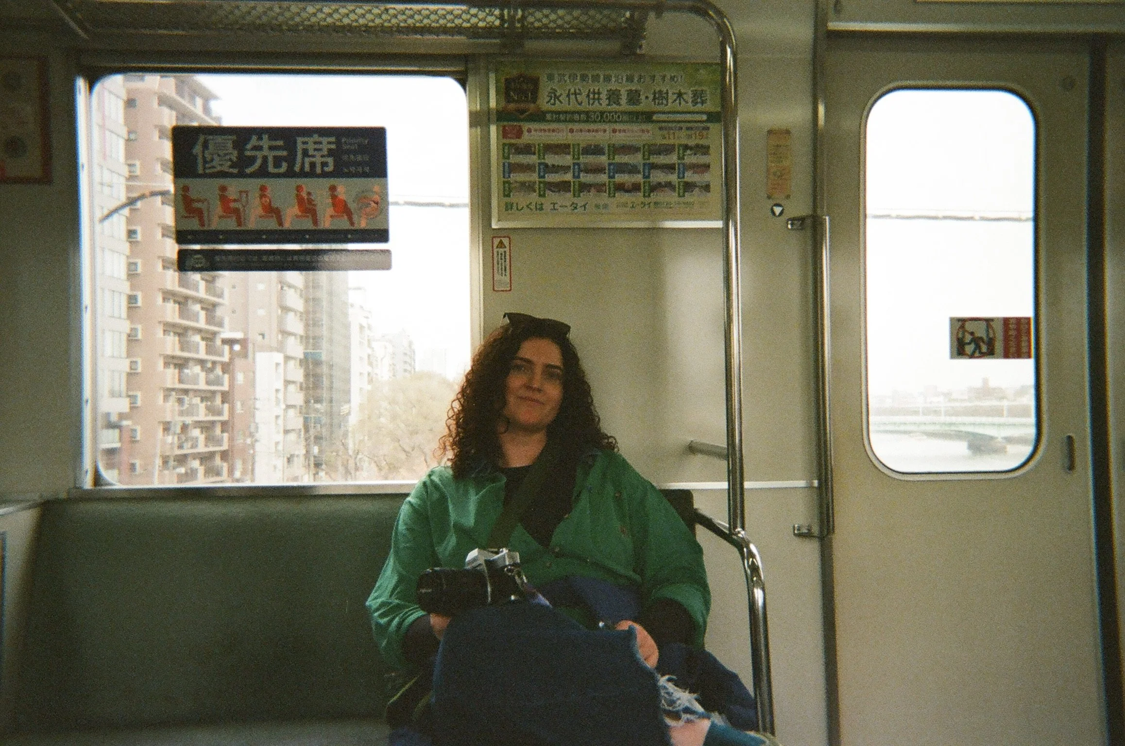 A film photographer, sitting inside a train, holding a film camera and smiling. The background shows an urban landscape with residential buildings through the train window.