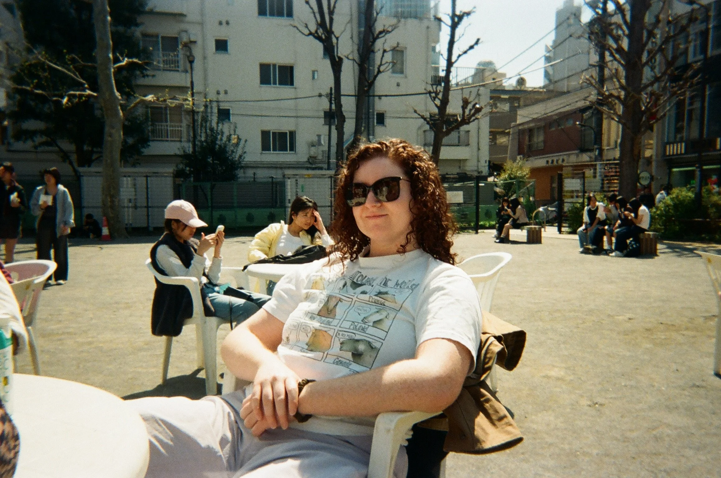 A woman with curly hair and sunglasses sitting outdoors in a park, smiling at the camera, with other people sitting and walking in the background.