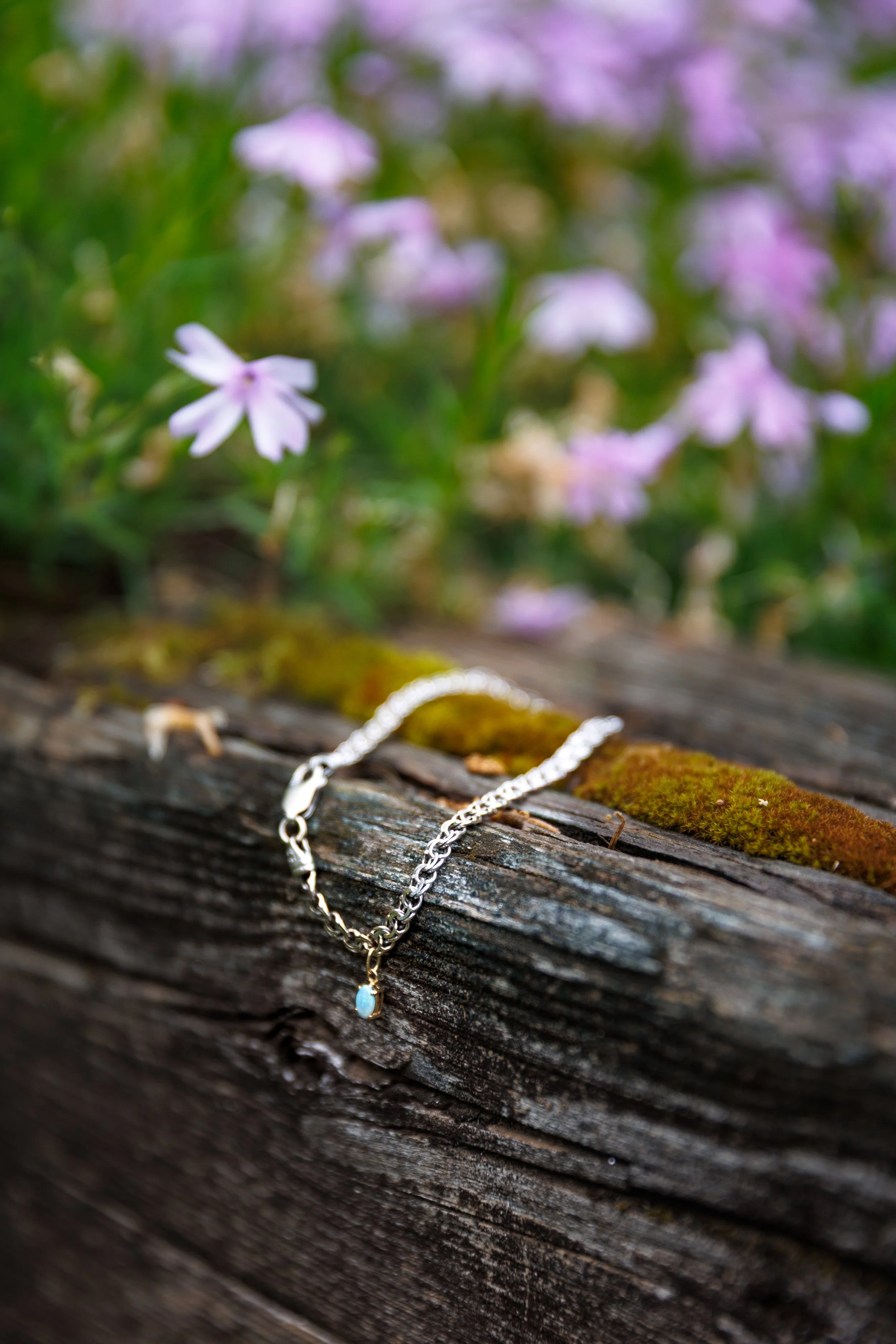 Silver necklace with a pendant resting on a weathered wooden log, with blurred purple flowers in the background.