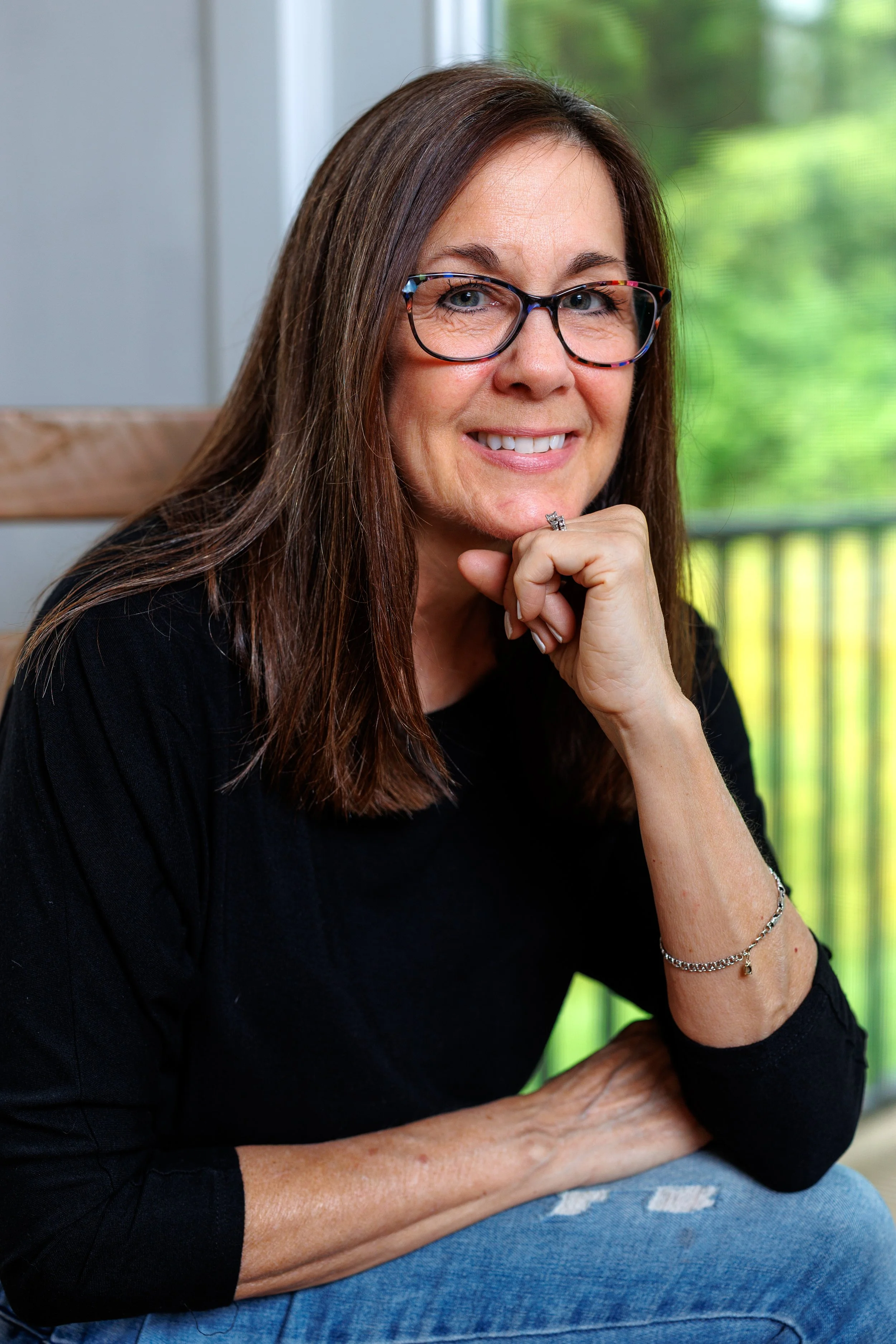 Marianne Heffernan smiling and resting her chin on her hand, sitting indoors near a window with a green view outside.