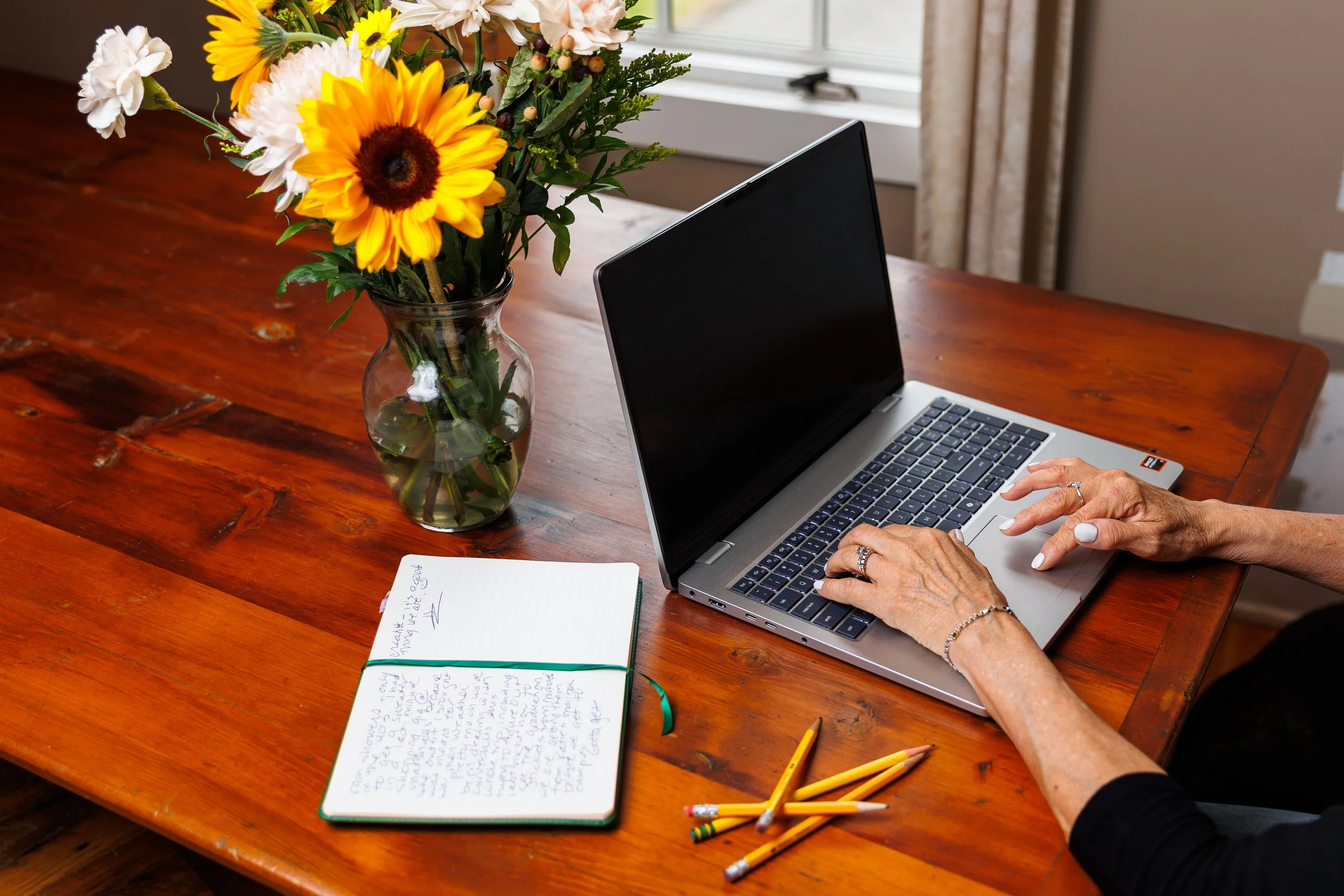 A woman working on a laptop at a wooden table with a bouquet of sunflowers and white flowers in a glass vase, a notebook with handwritten notes, and several yellow pencils nearby.