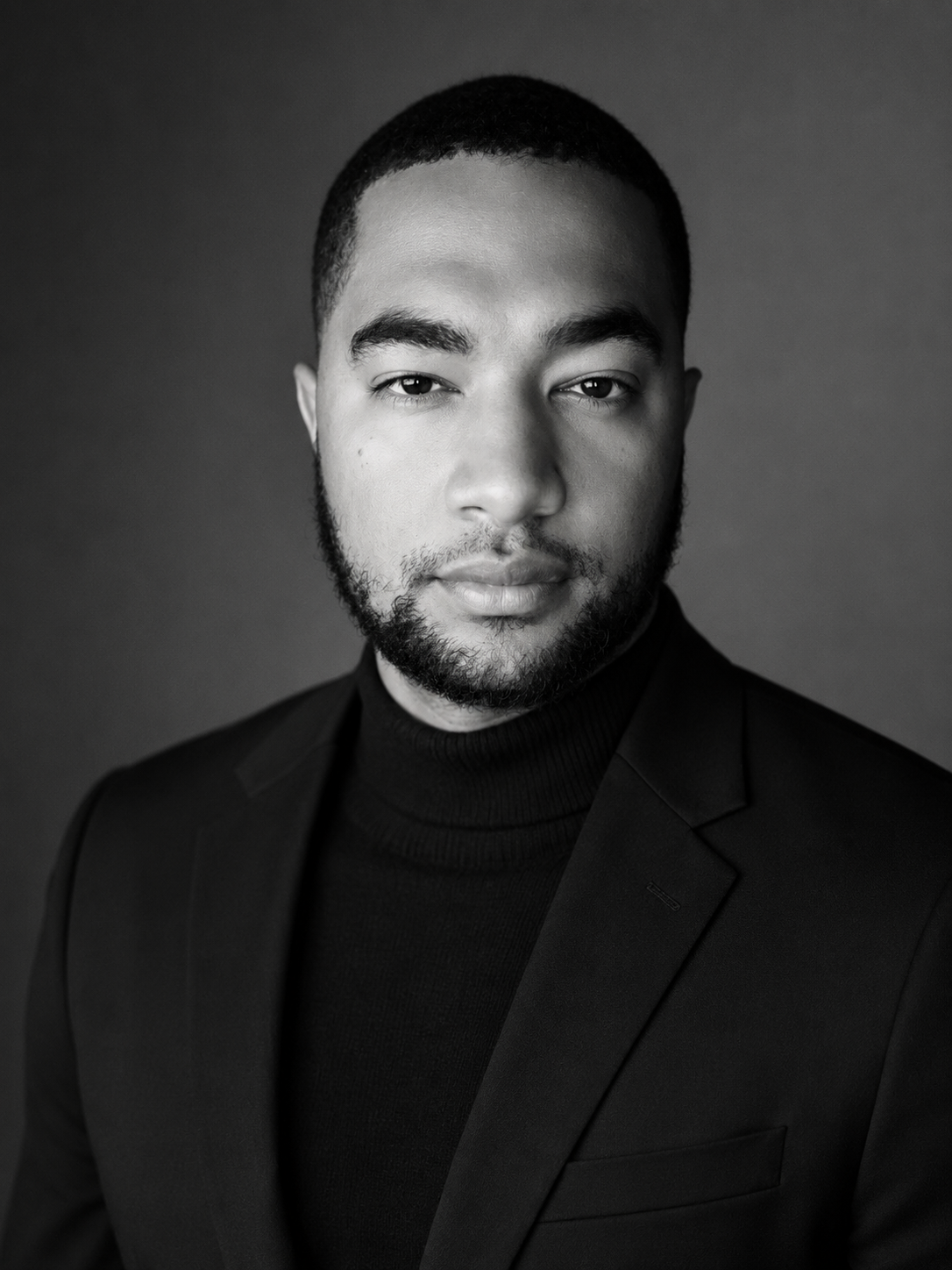 Black and white portrait of a young man with short hair and a beard, wearing a dark blazer and turtleneck sweater, looking directly at the camera against a plain background.