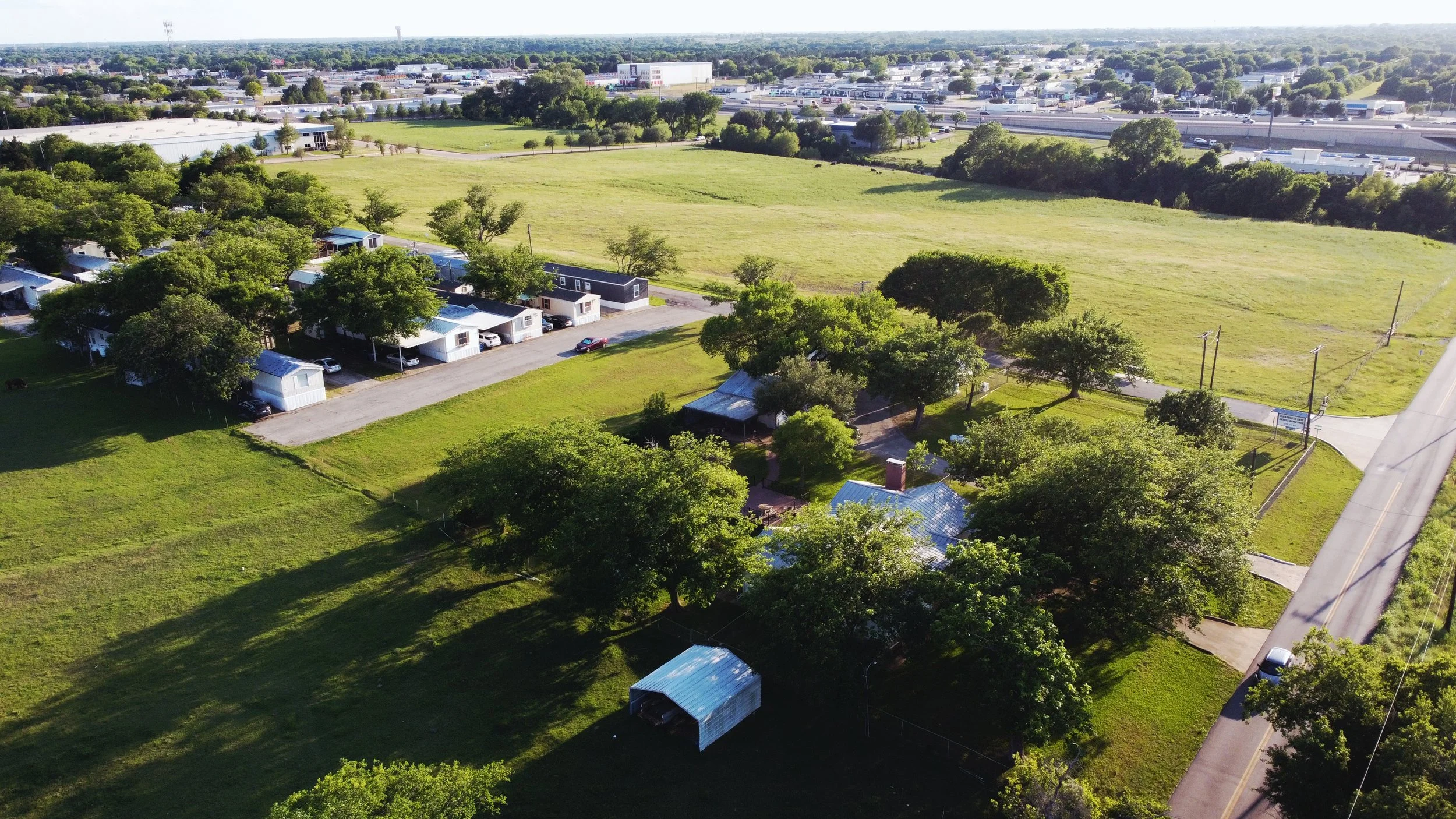 Aerial view of a residential neighborhood with small houses, green trees, and grassy fields adjacent to a busy commercial area with shopping centers and roads in the background.