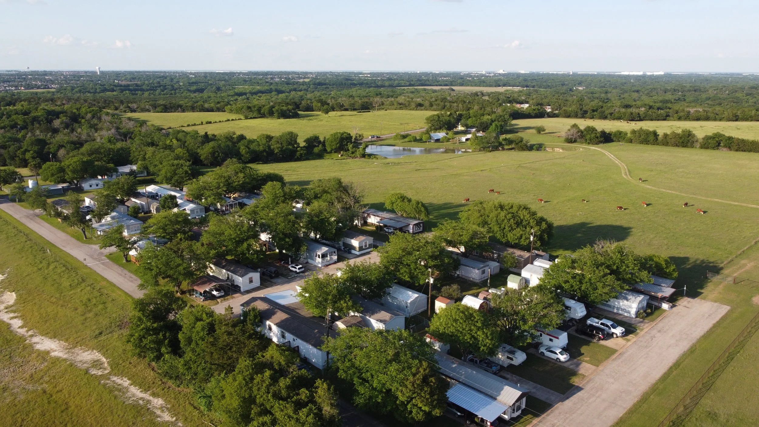 An aerial view of a rural landscape with green fields, scattered trees, a pond, and a small neighborhood with mobile homes and cars.
