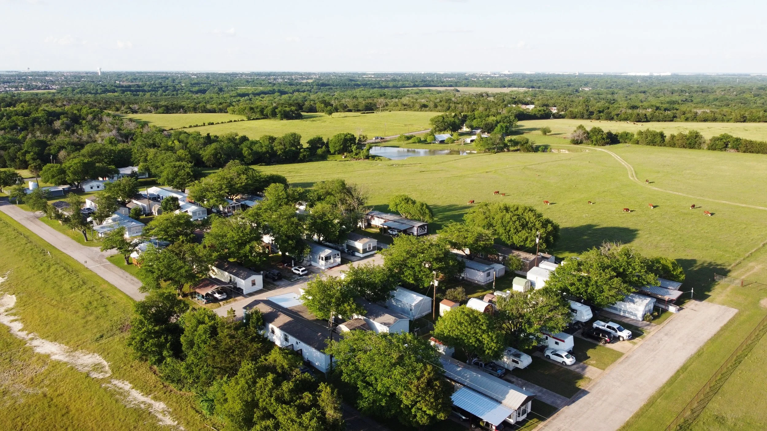 Aerial view of a rural area with mobile homes and RVs along a road, green fields, trees, a pond, and distant city skyline.