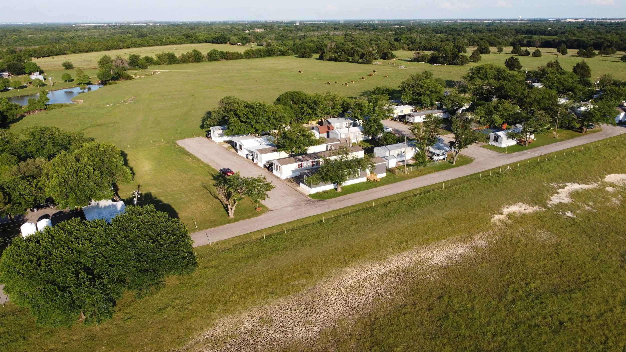 Aerial view of a small residential area with mobile homes surrounded by green trees and open fields, with a pond nearby and a distant skyline.