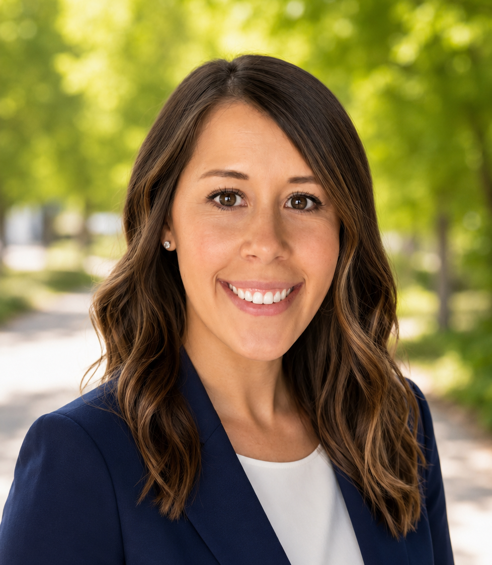 A woman with brown hair styled in loose waves, wearing a navy blazer and a white blouse, smiling outdoors with a blurred green leafy background.