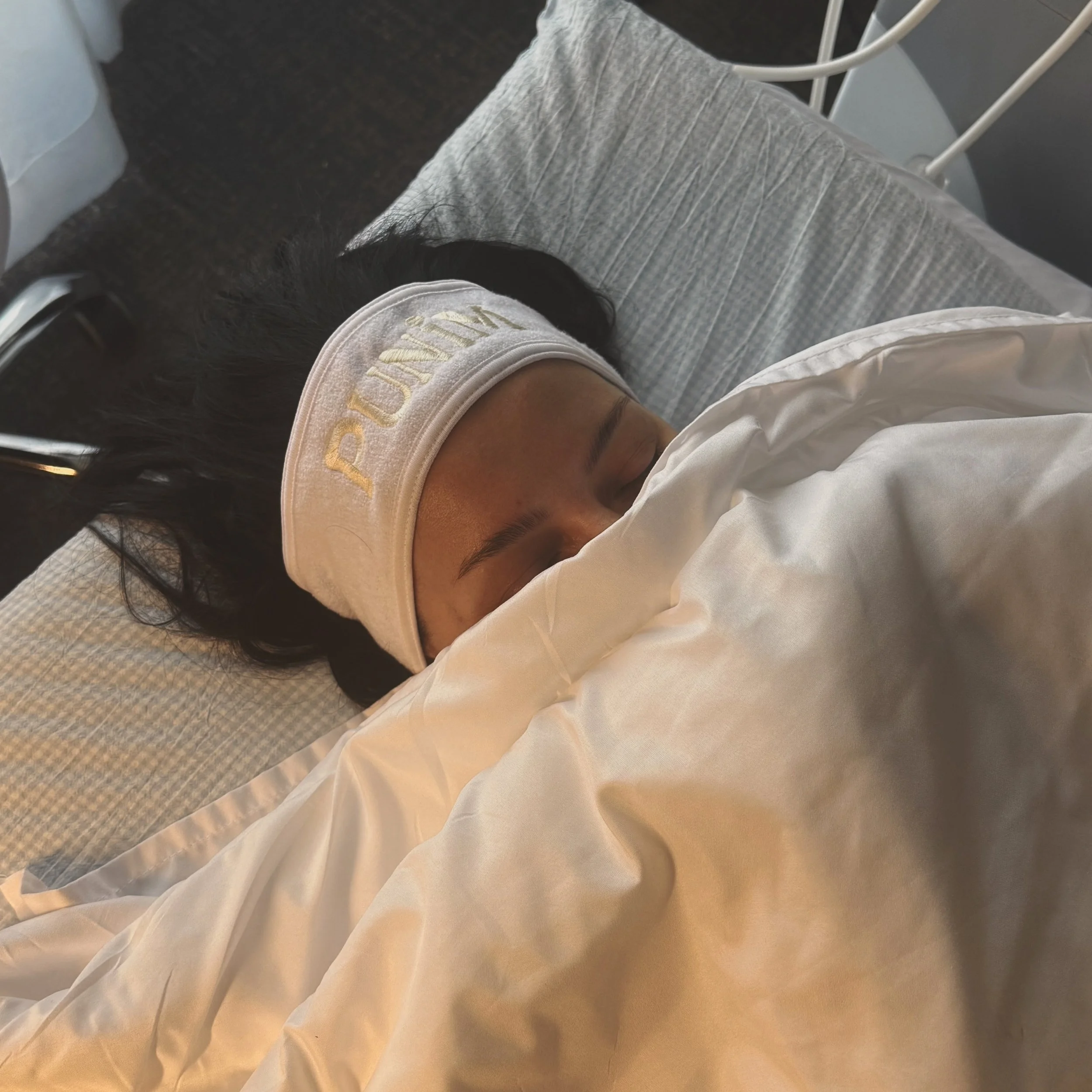 A person lying on a bed wearing a white headband with gold embroidery, with closed eyes and long dark hair, covered with a white blanket or sheet. There is a gray upholstered headboard and a dark floor in the background.