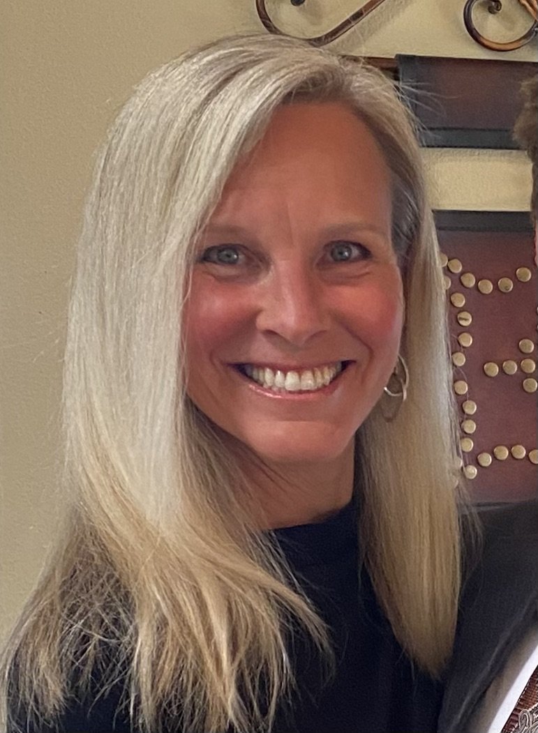 Tammy Wheeler, smiling and wearing a black top, standing indoors with a decorative background.