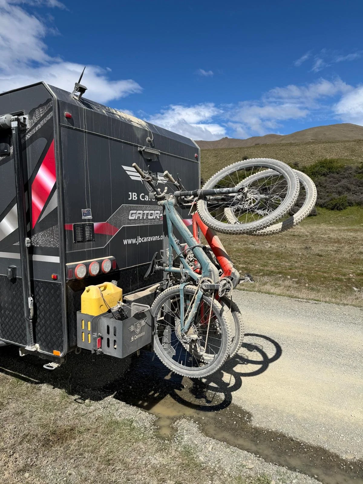 A black caravan with EziLift holding two electric mountain bikes is parked outdoors on a dirt road with a grassy landscape and hills in the background.