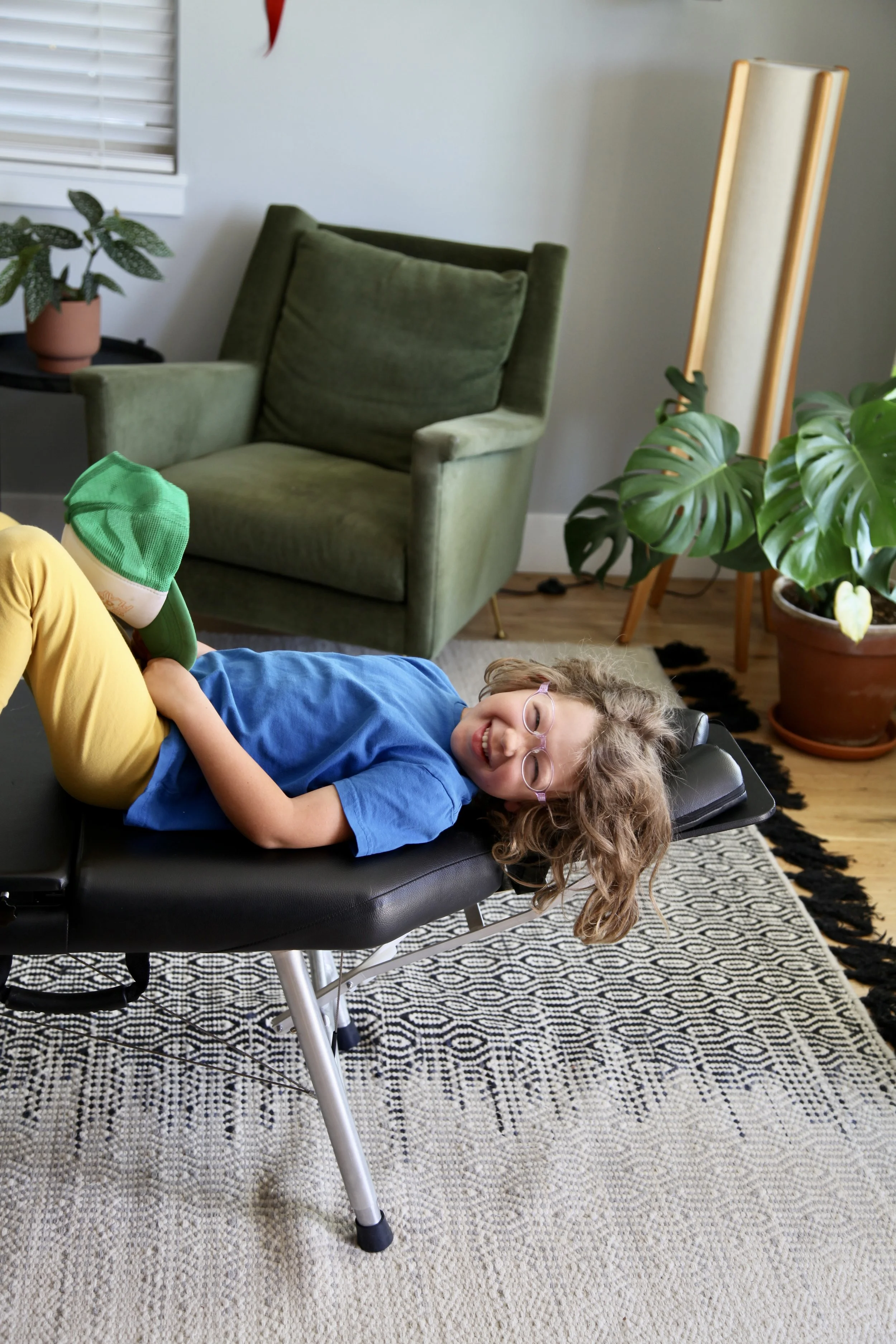 A young boy with curly hair and glasses lying on his back on a chiropractic table, smiling at the camera. The background shows a room with green and beige furniture, potted plants, a black and white patterned rug, and a window with blinds.