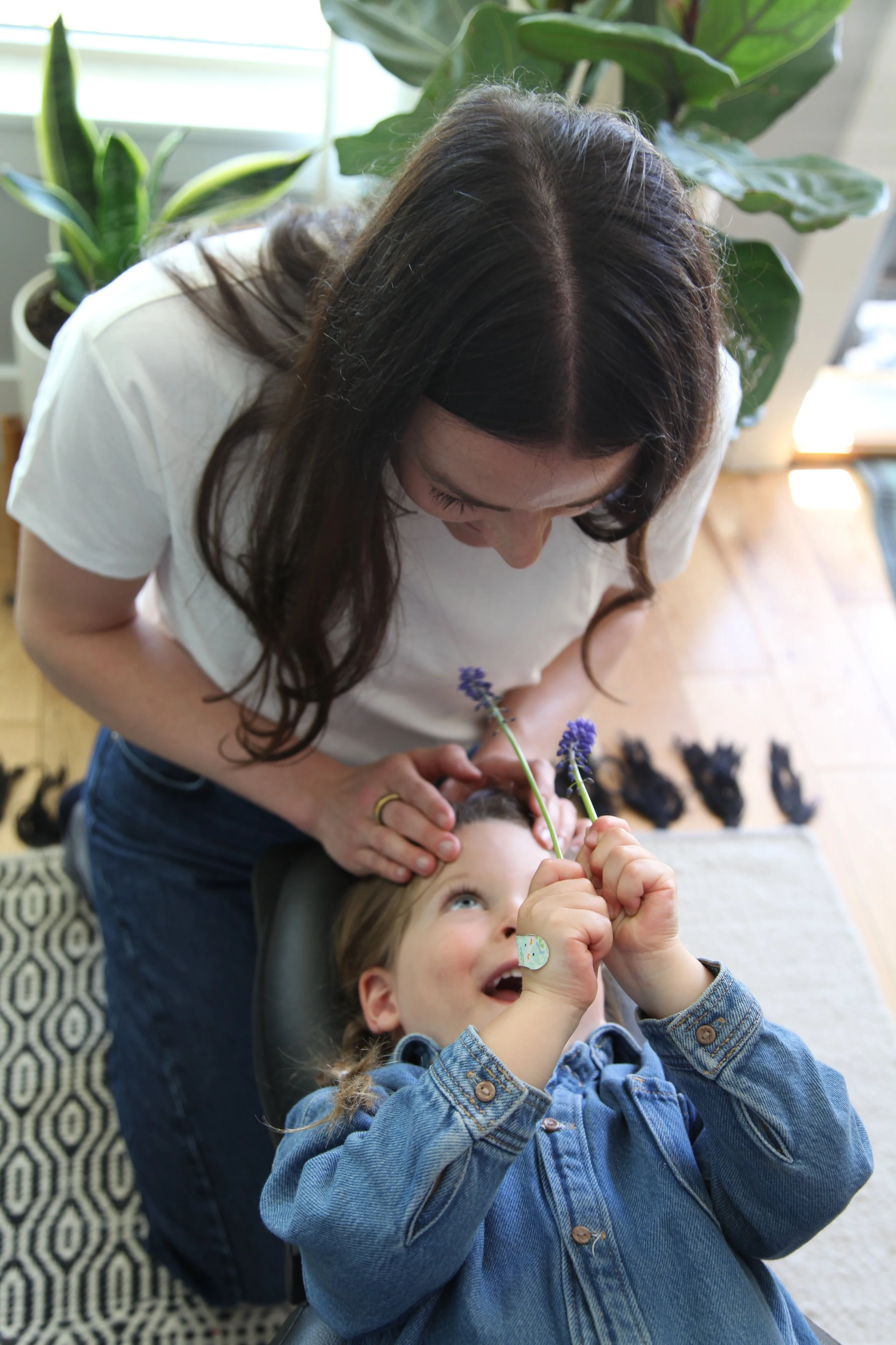 A woman is adjusting a child, while the boy is playing with flowers. The boy is smiling and holding flowers in his hands. The scene is indoors, with large green plants in the background and shoes visible on the floor.