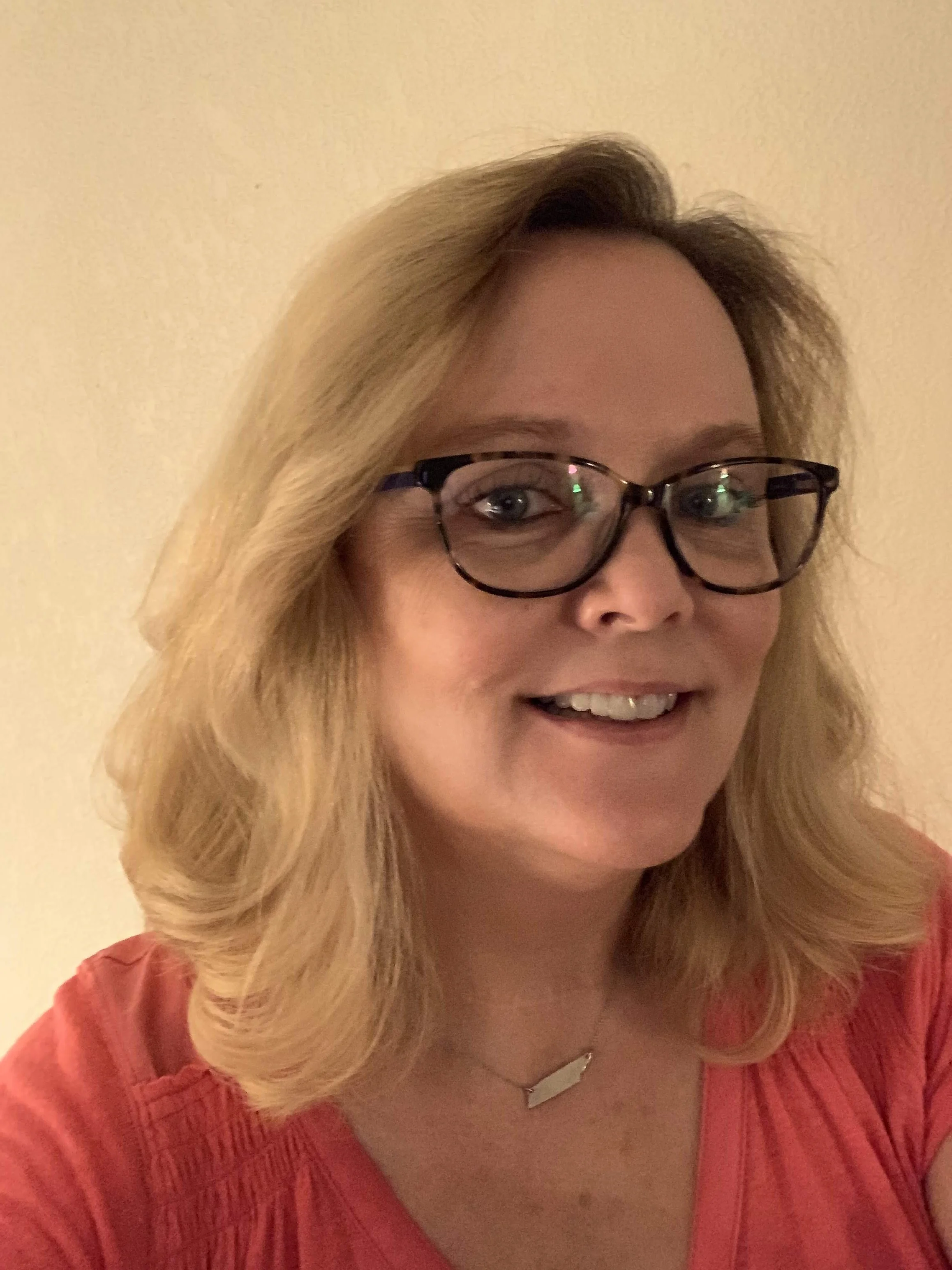 A woman with blonde hair and glasses smiling, wearing a red top and a silver necklace, against a beige background.