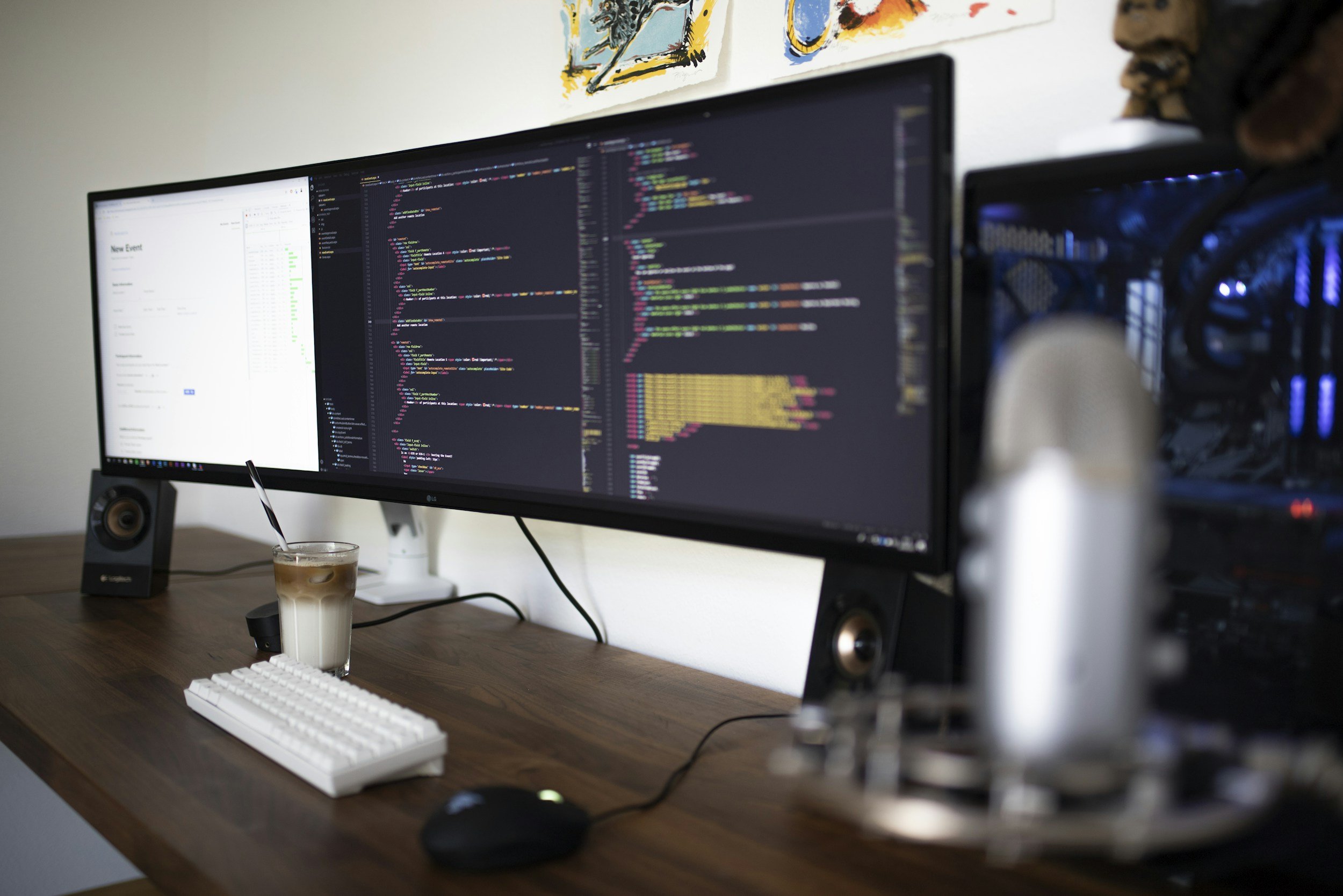 A dual-monitor computer setup on a wooden desk, with a white keyboard and mouse, a glass of iced coffee, and a speaker. The monitors display colorful lines of code, and a person is working at the desk, with part of their face and curly hair visible.
