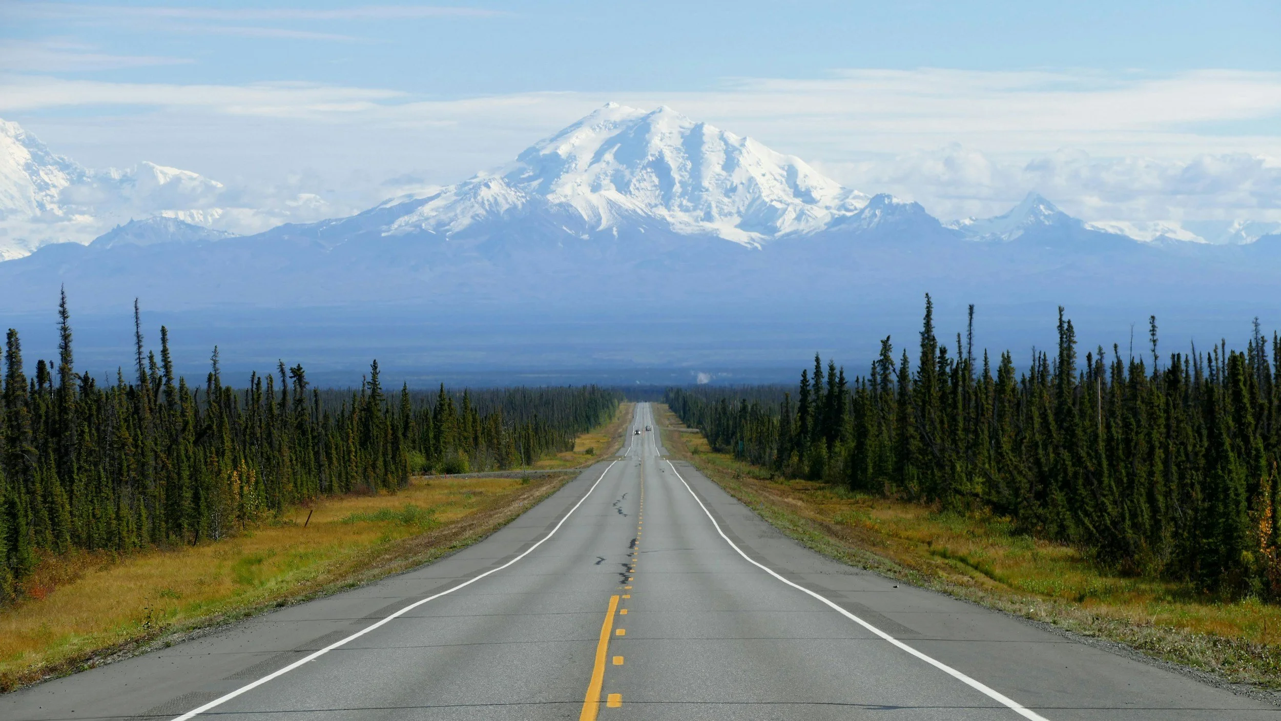 A two-lane highway leading towards a large snow-capped mountain in the distance, flanked by dense green trees on both sides under a partly cloudy sky.