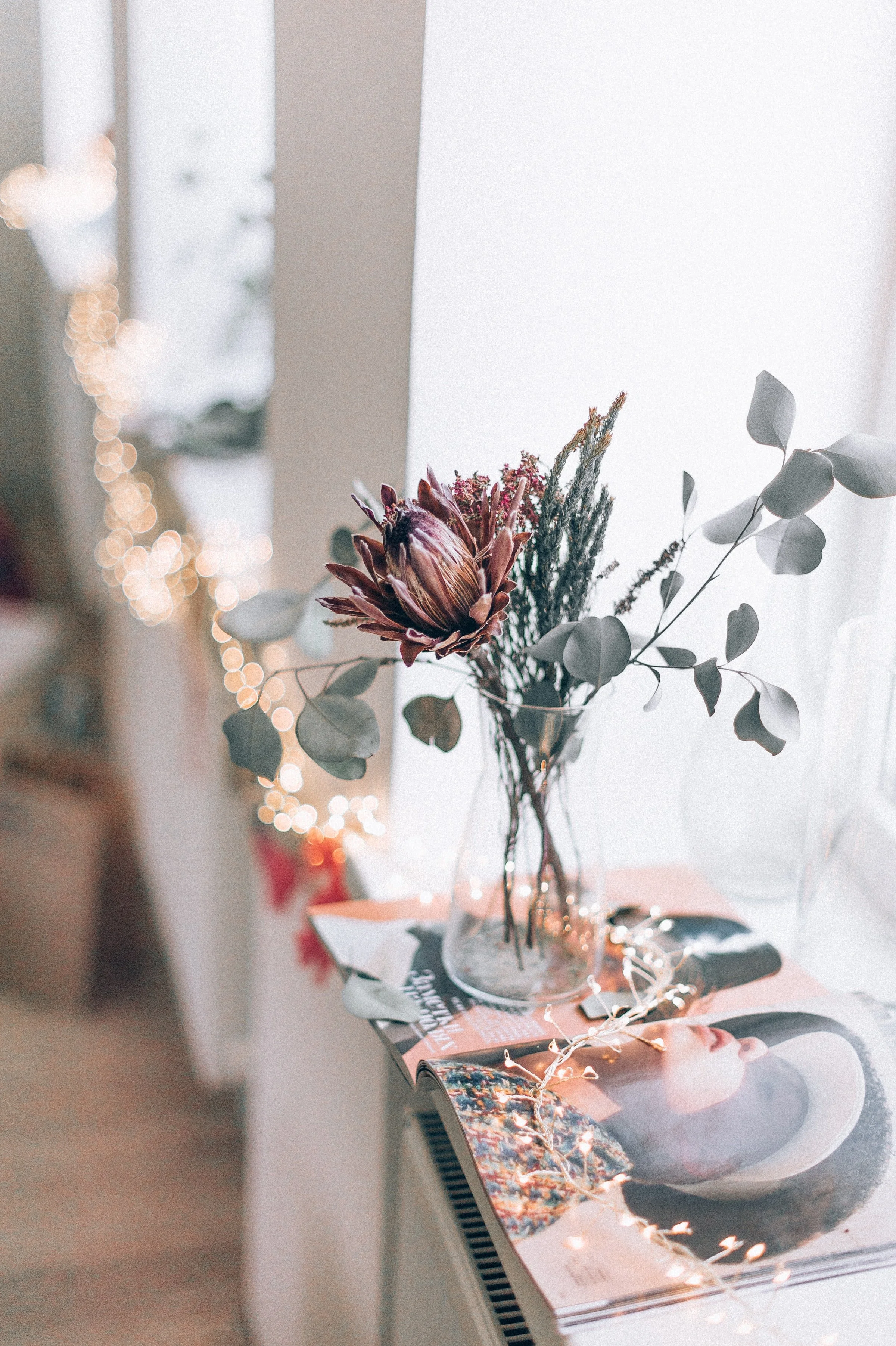 A bouquet of dried flowers with leaves in a glass vase placed on a table with open magazines. The scene is lit softly, with bokeh lights in the background.