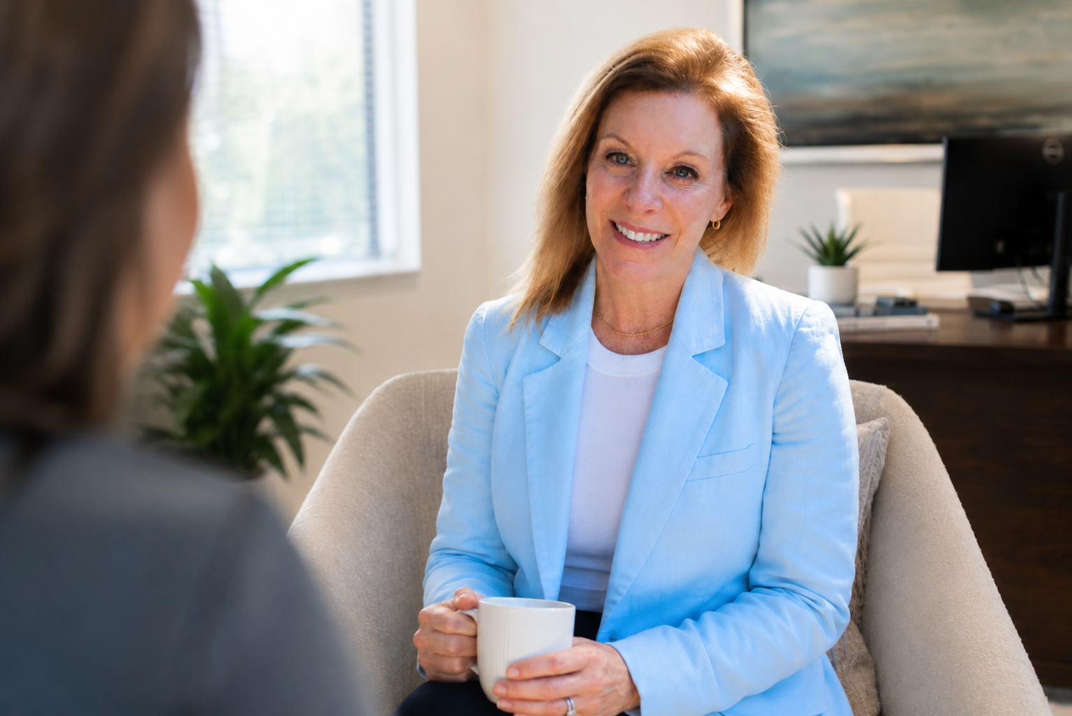 A woman with red hair wearing a light blue blazer and holding a white mug, smiling during a conversation in a cozy office or living room setting.