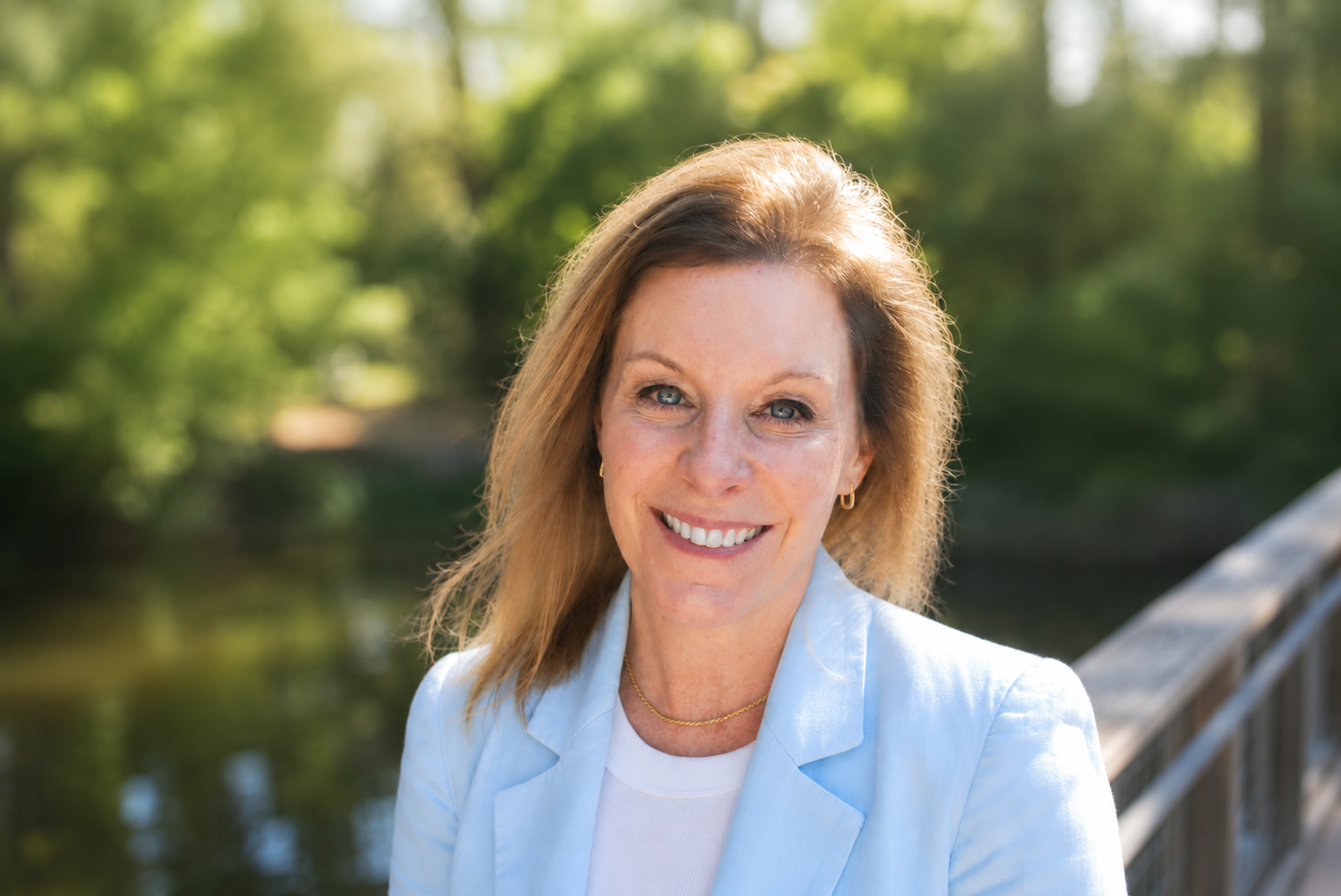 A woman with blonde hair smiling outdoors on a wooden bridge, with green trees and water in the background.