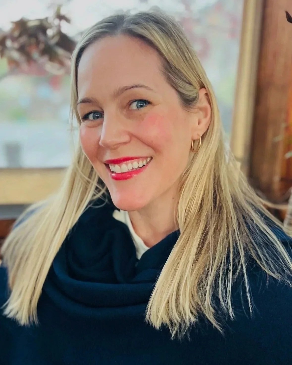 A woman with long blonde hair and blue eyes is smiling at the camera. She is wearing gold hoop earrings, red lipstick, and a black top with a white shirt underneath. The background appears to be indoors with a window and some plants.