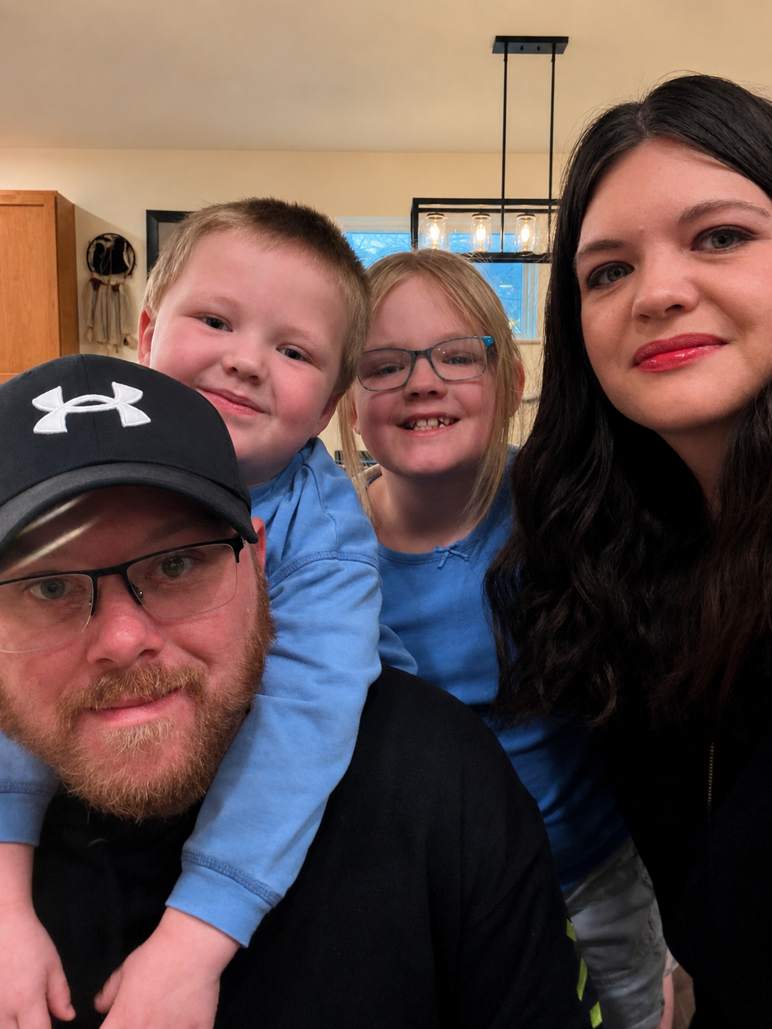 A family selfie featuring a man with a beard and glasses wearing a black Under Armour cap, a woman with long dark hair and red lipstick, a young girl with blonde hair and glasses, and a young boy with blonde hair, all smiling and close together indoors.