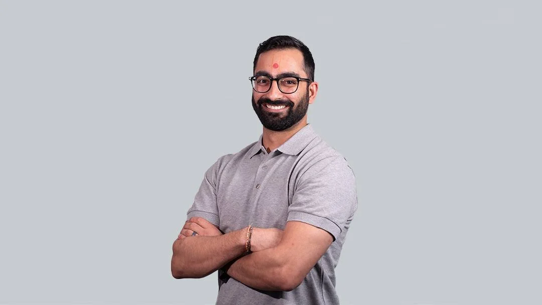 A smiling man with glasses, a beard, and a tilak on his forehead, standing with arms crossed against a plain light gray background.