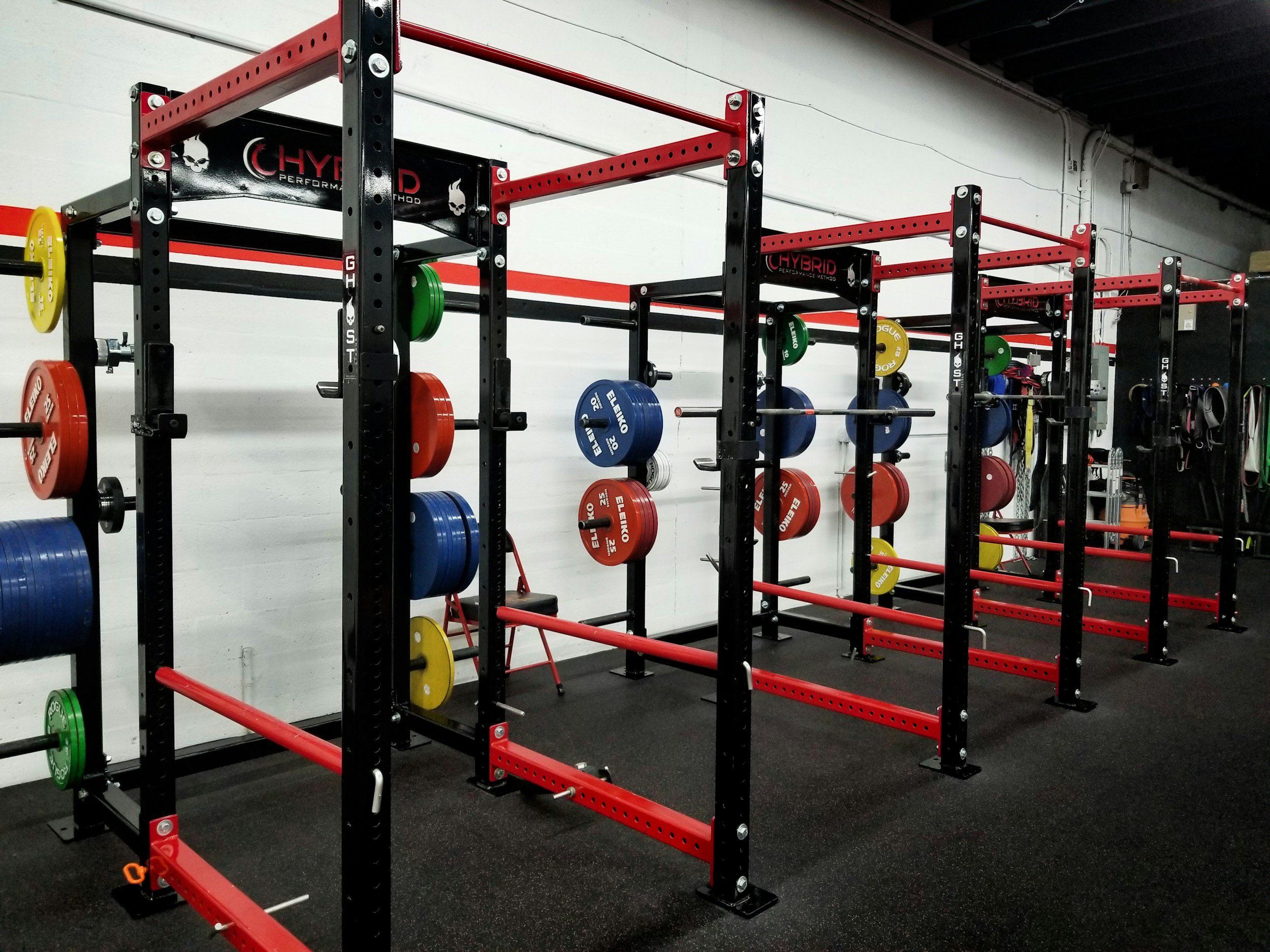 Inside a gym, black and red power racks with colored bumper plates, resistance bands, and workout equipment organized in the background.
