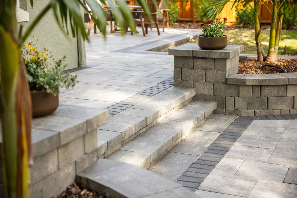 Newly paved backyard patio with stairs, potted plants, and garden beds surrounded by lush greenery and trees.