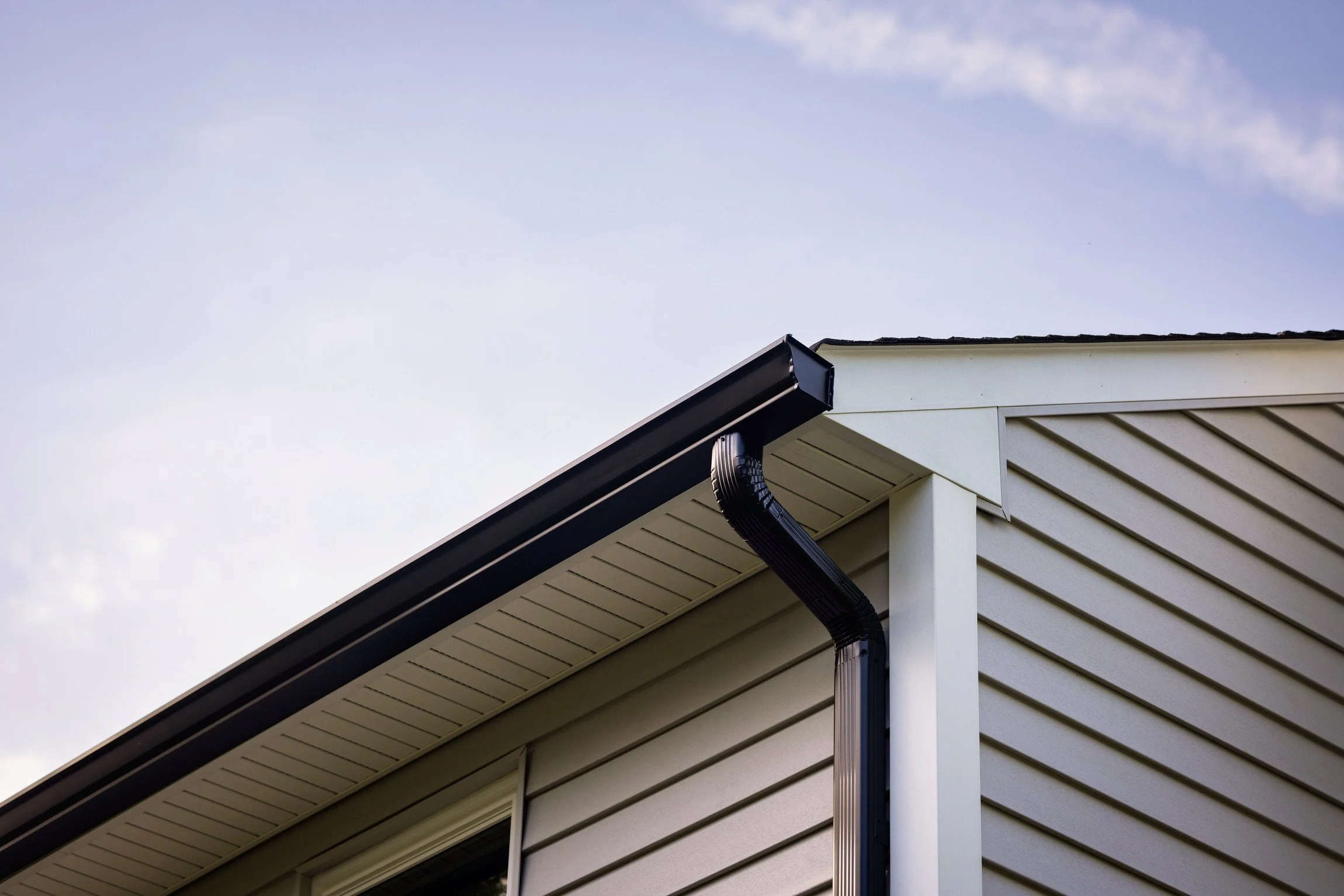 A close-up view of the corner of a house showing beige horizontal siding, a black gutter, and a blue sky with some clouds.