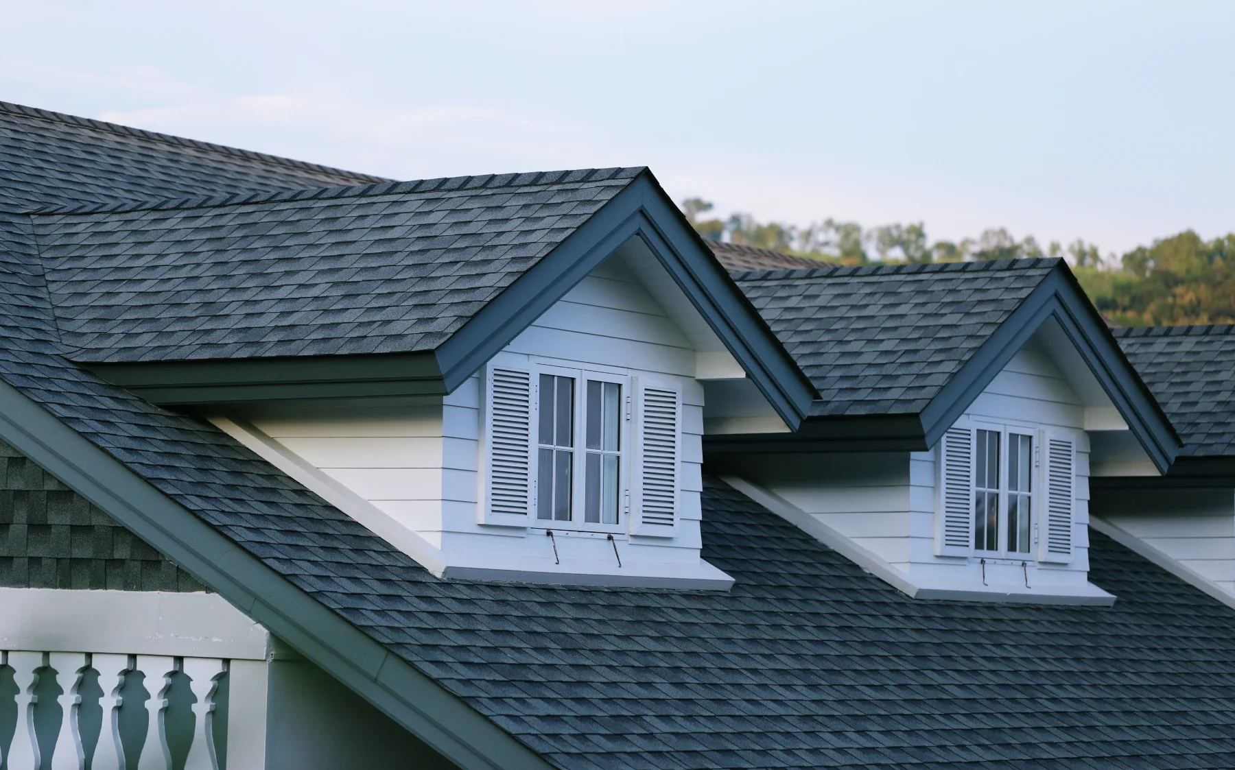 Close-up of a house roof with gray shingles and white dormer windows with shutters, green trees in the background.