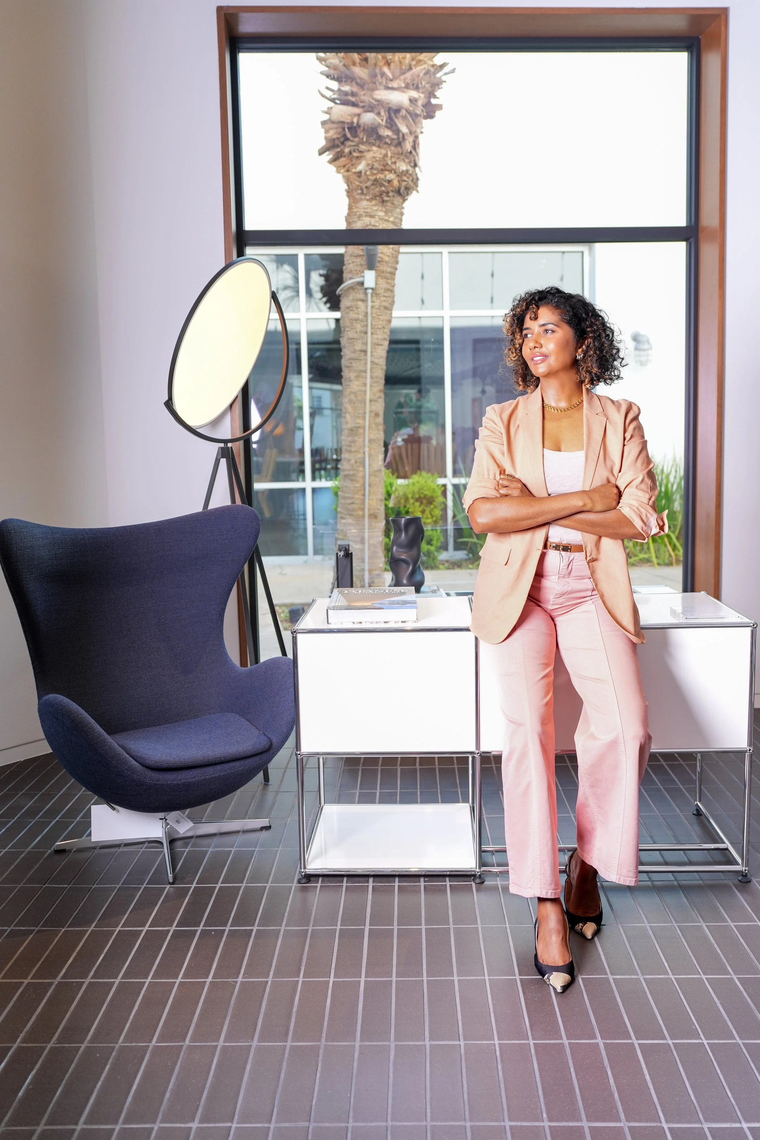 A woman with curly hair wearing a peach blazer and pants stands with her arms crossed inside a modern office lobby, with a large window showing a palm tree outside. There is a dark blue chair, a white side table with books, and a standing mirror in the interior.